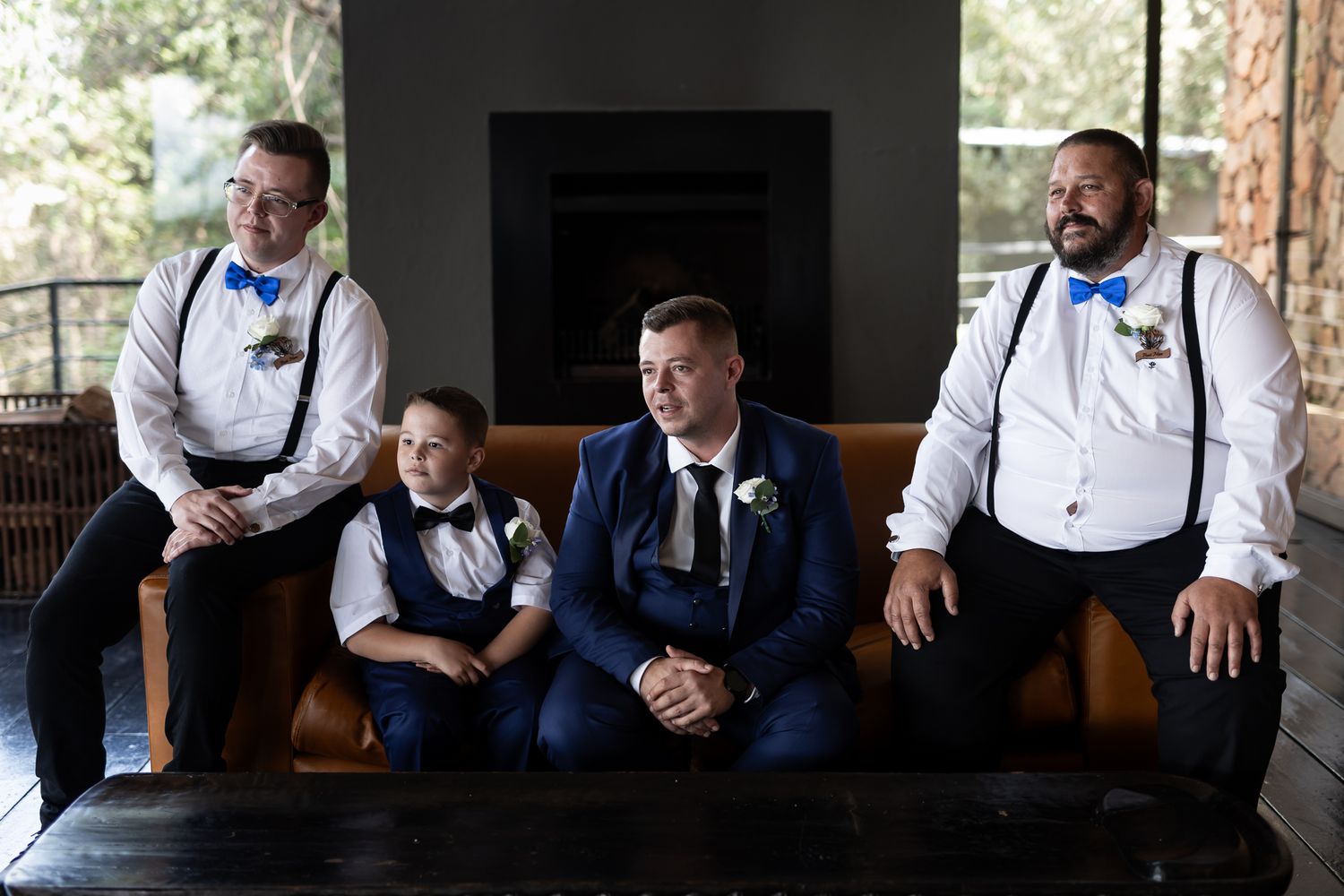 A wedding group sits casually on a dark leather couch during formal portraits.