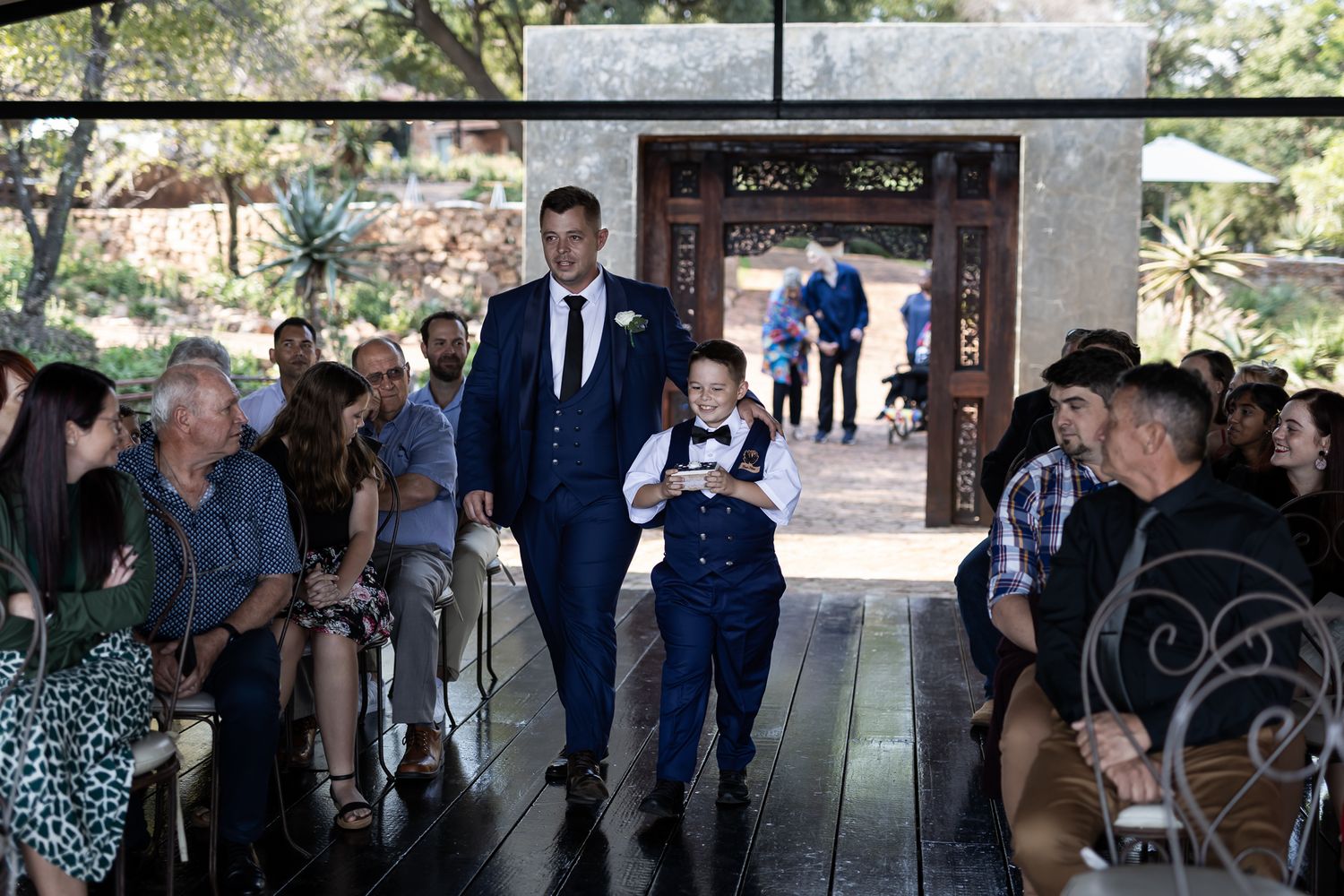 Wedding party members walk down a covered outdoor aisle lined with seated guests.