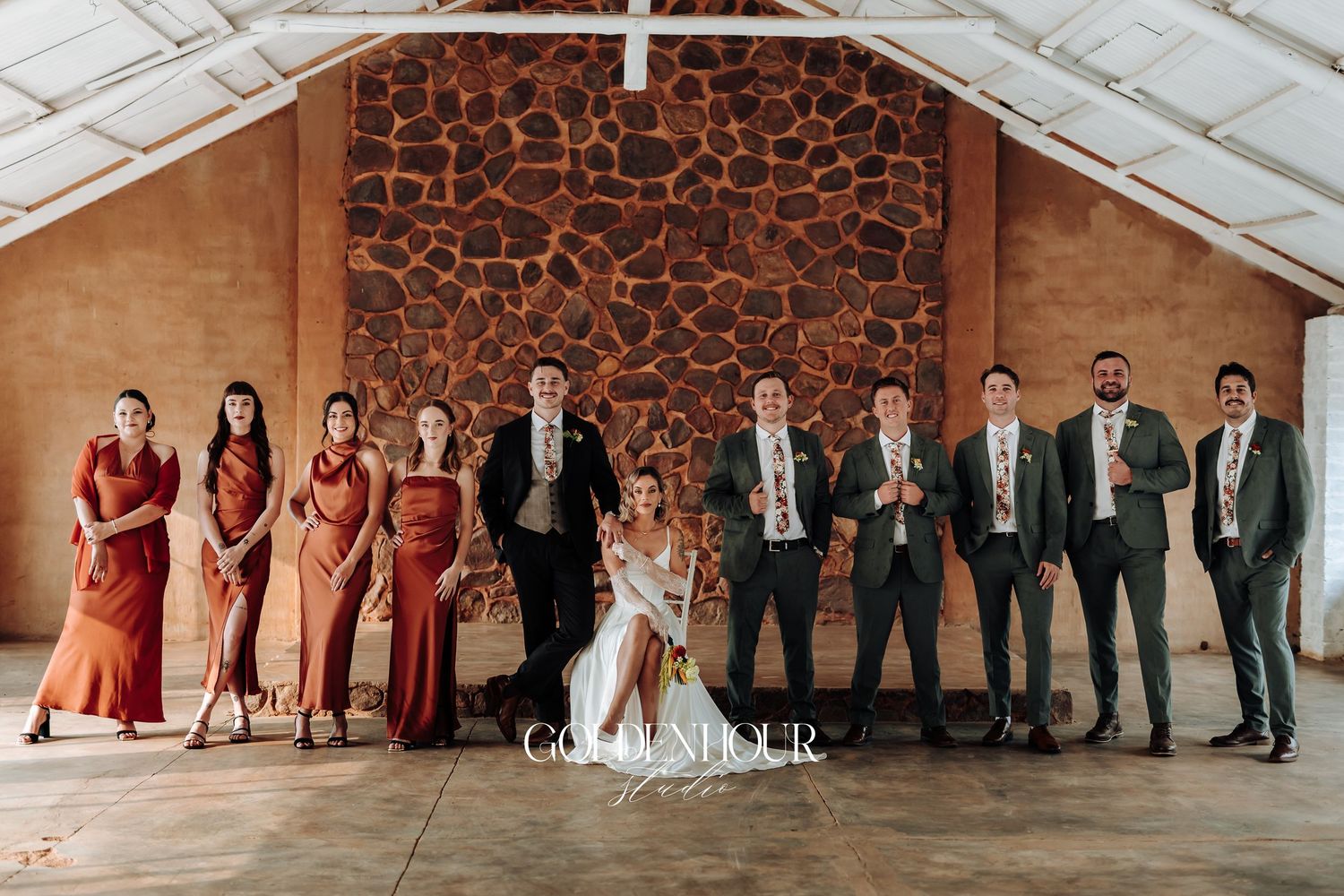 Wedding party lineup showing bridesmaids in burgundy and groomsmen in black suits against rustic wall.