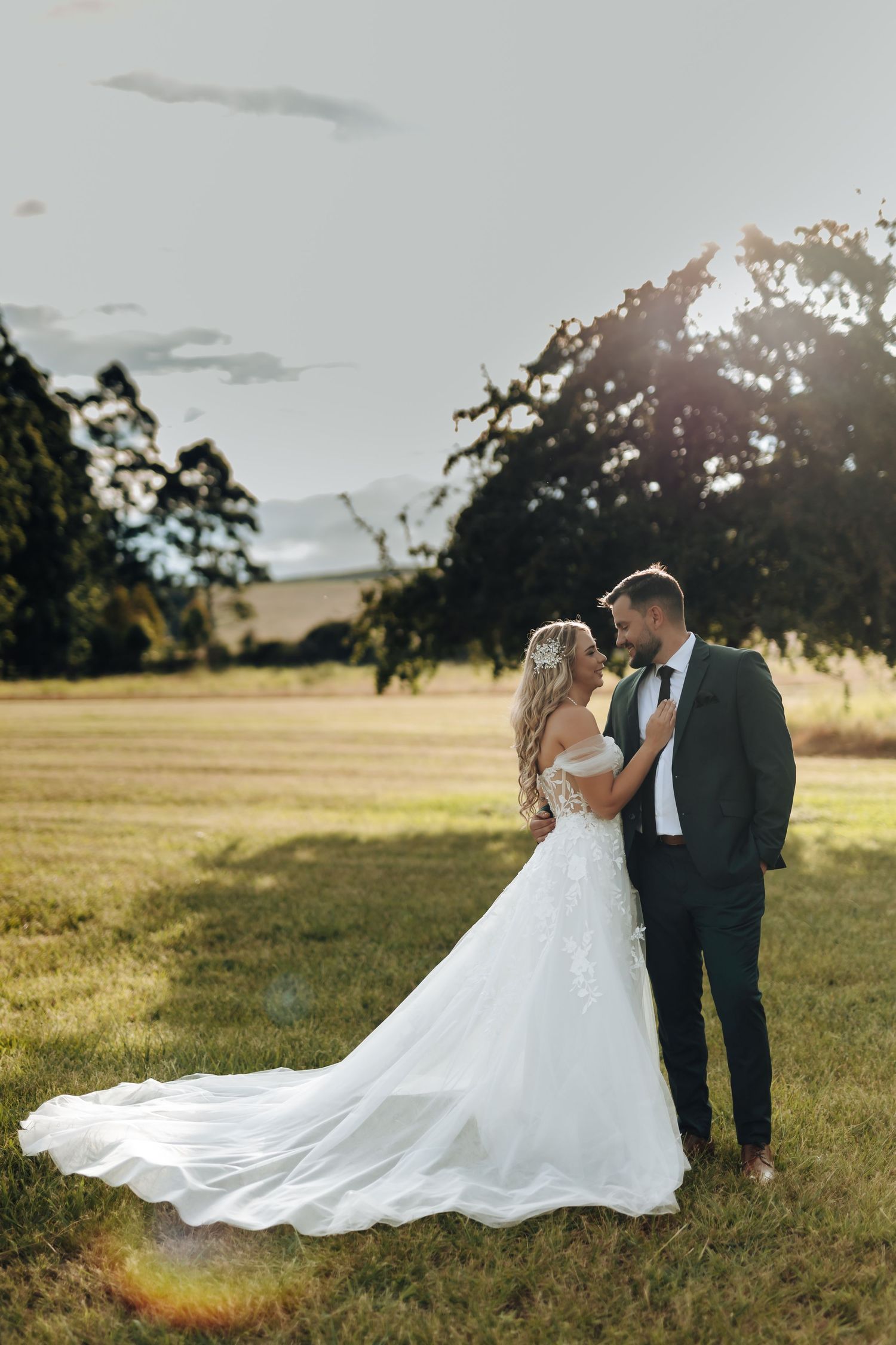 A romantic wedding couple embraces in a sunlit meadow at golden hour with a flowing white dress trailing behind them.