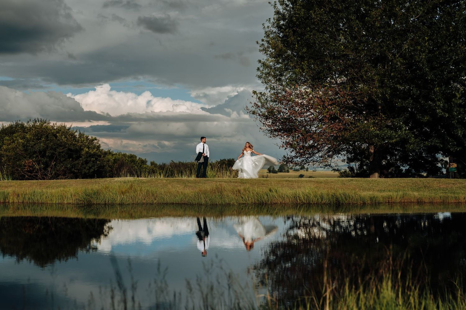 Couple walking along a serene lake shoreline with dramatic cloudy sky reflected in still waters at sunset.