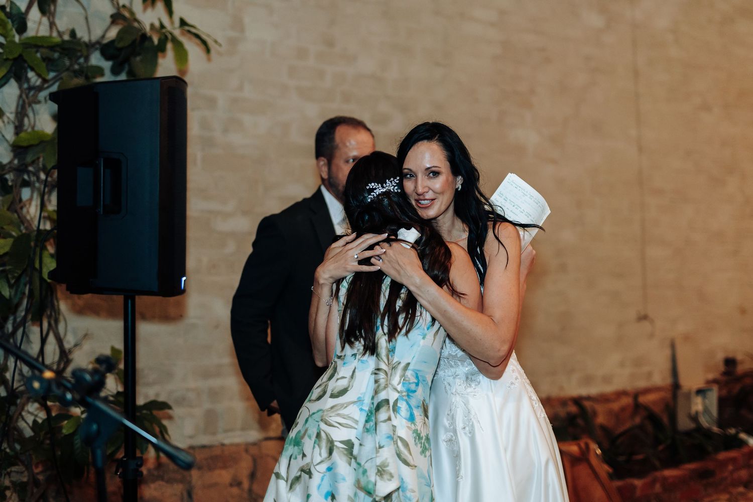 Bride in white gown hugging guest at wedding reception with man in suit and greenery in background.