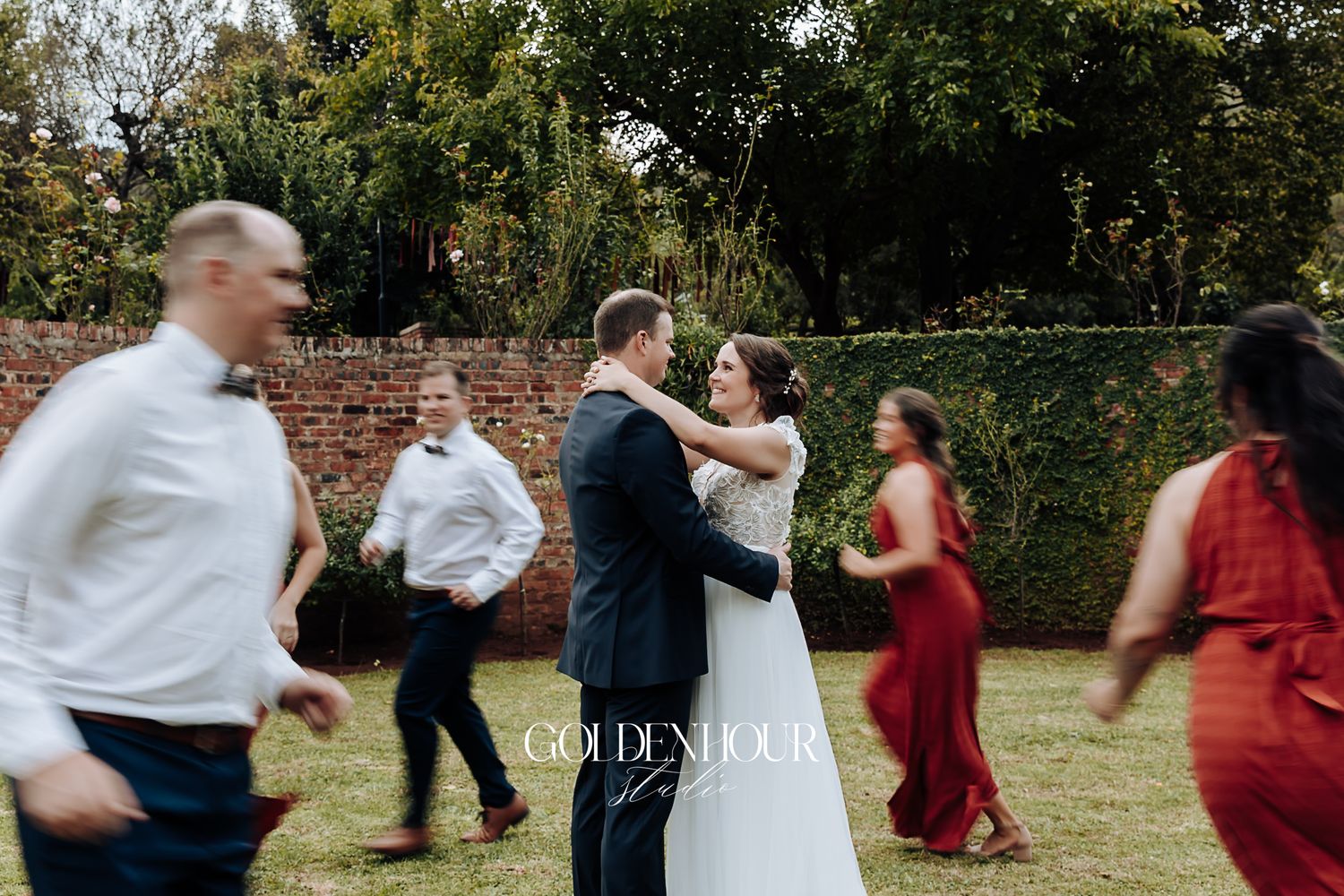 Bride and groom share first dance surrounded by blurred guests on green lawn at reception.