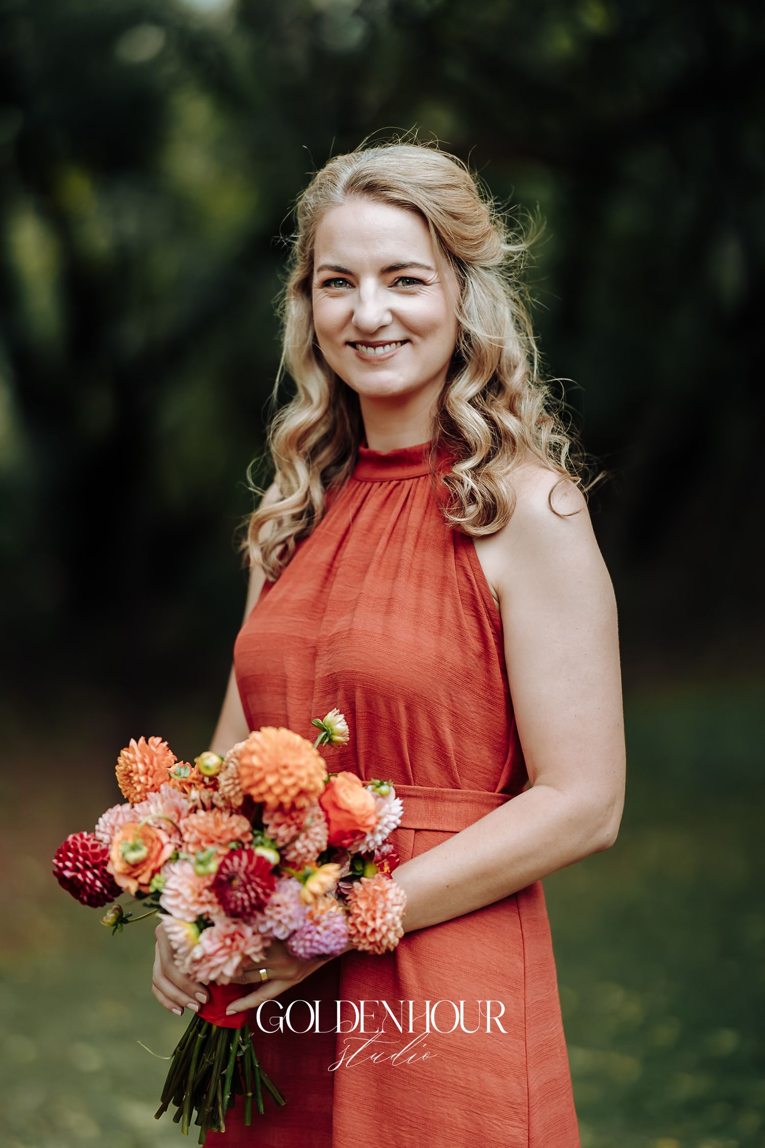 Blonde bridesmaid in terracotta halter dress holding a peach and coral dahlia bouquet outdoors.