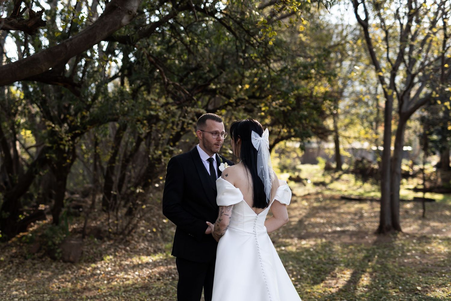 A couple in formal wedding attire embraces in a sunlit wooded area during autumn.