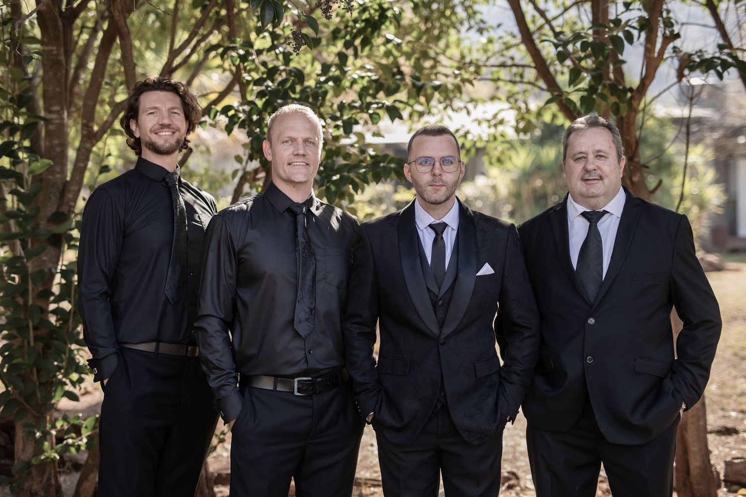 Group of four men in dark suits and ties posing together in a garden setting.