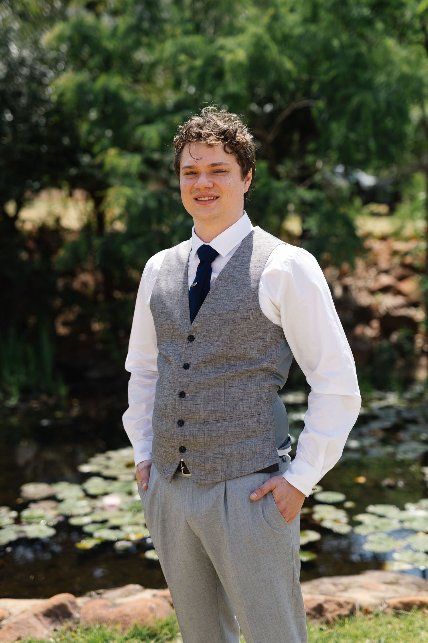 Person in formal attire poses outdoors by a pond with lily pads in a garden setting.