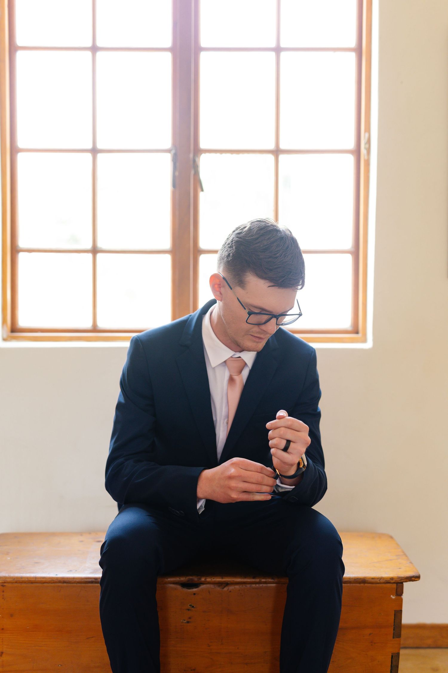 A person in a black suit and glasses sits on a wooden bench in a bright room with large windows.