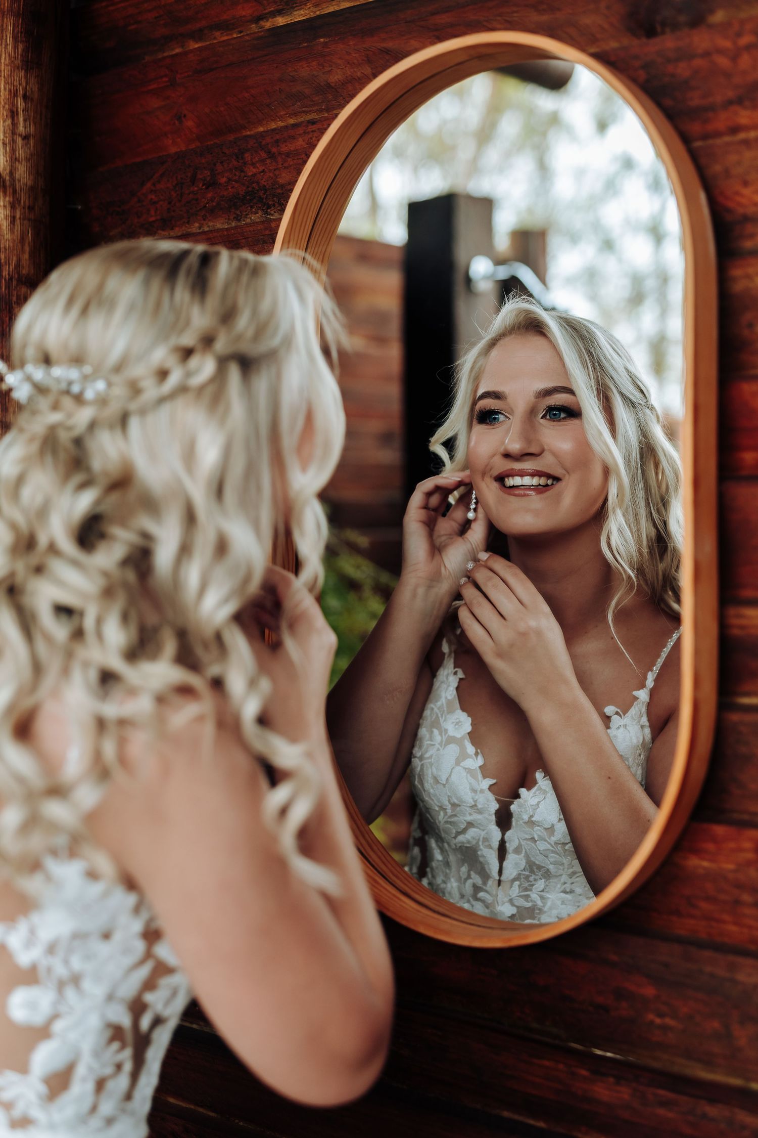 A bride in a lace wedding dress puts on earrings while looking at her reflection in an oval mirror mounted on wood.