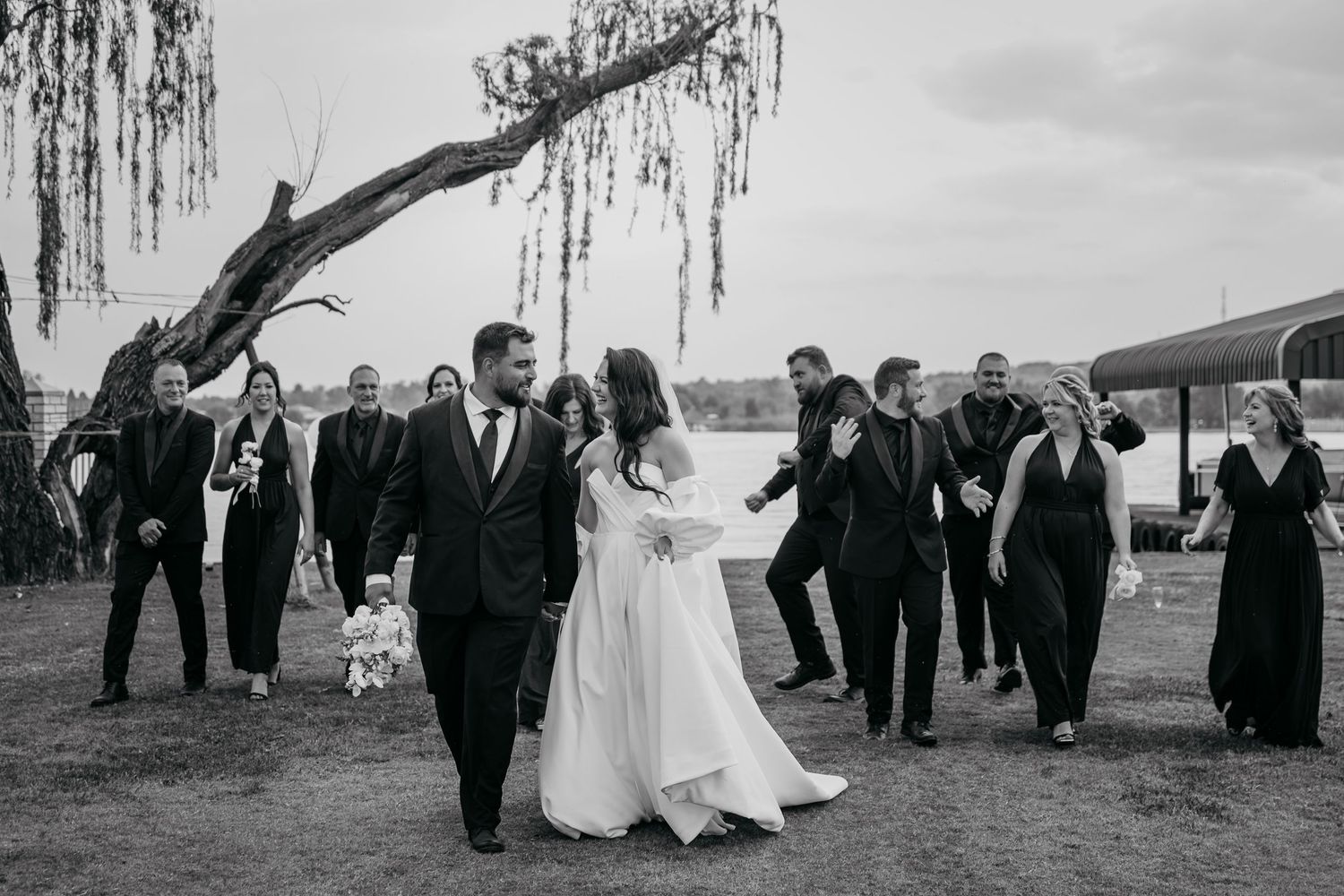 A wedding party poses together under a weeping willow tree near a waterfront on an overcast day.
