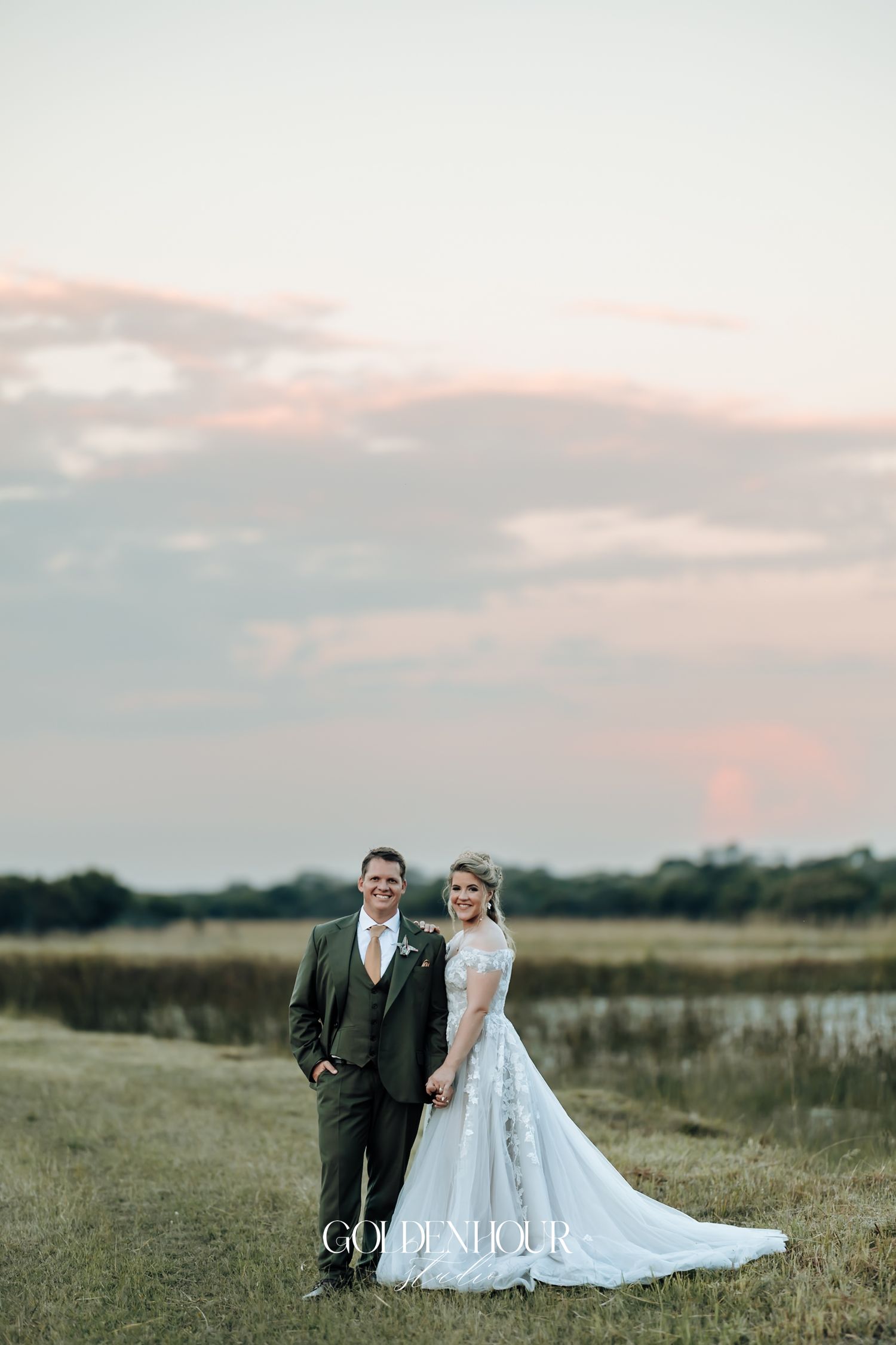 Bride and groom holding hands in an open field at sunset with a pastel pink sky backdrop.
