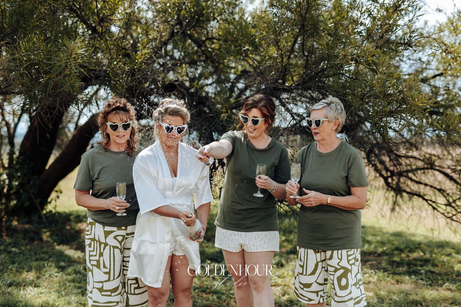 Bride in white robe with three bridesmaids in olive green shirts enjoying champagne outdoors under a tree.
