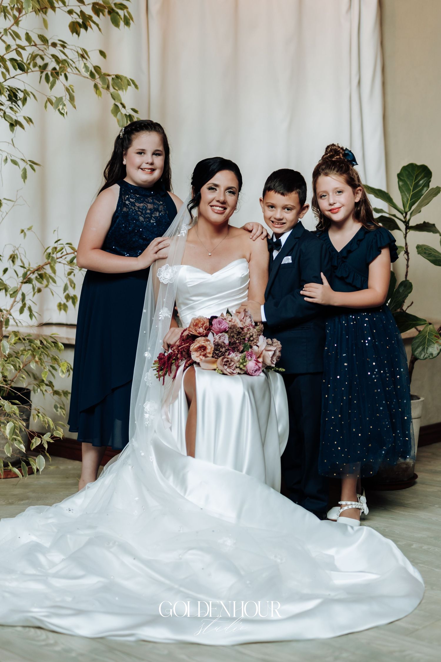 Smiling bride in white satin gown poses with three children in navy formal attire holding a floral bouquet.