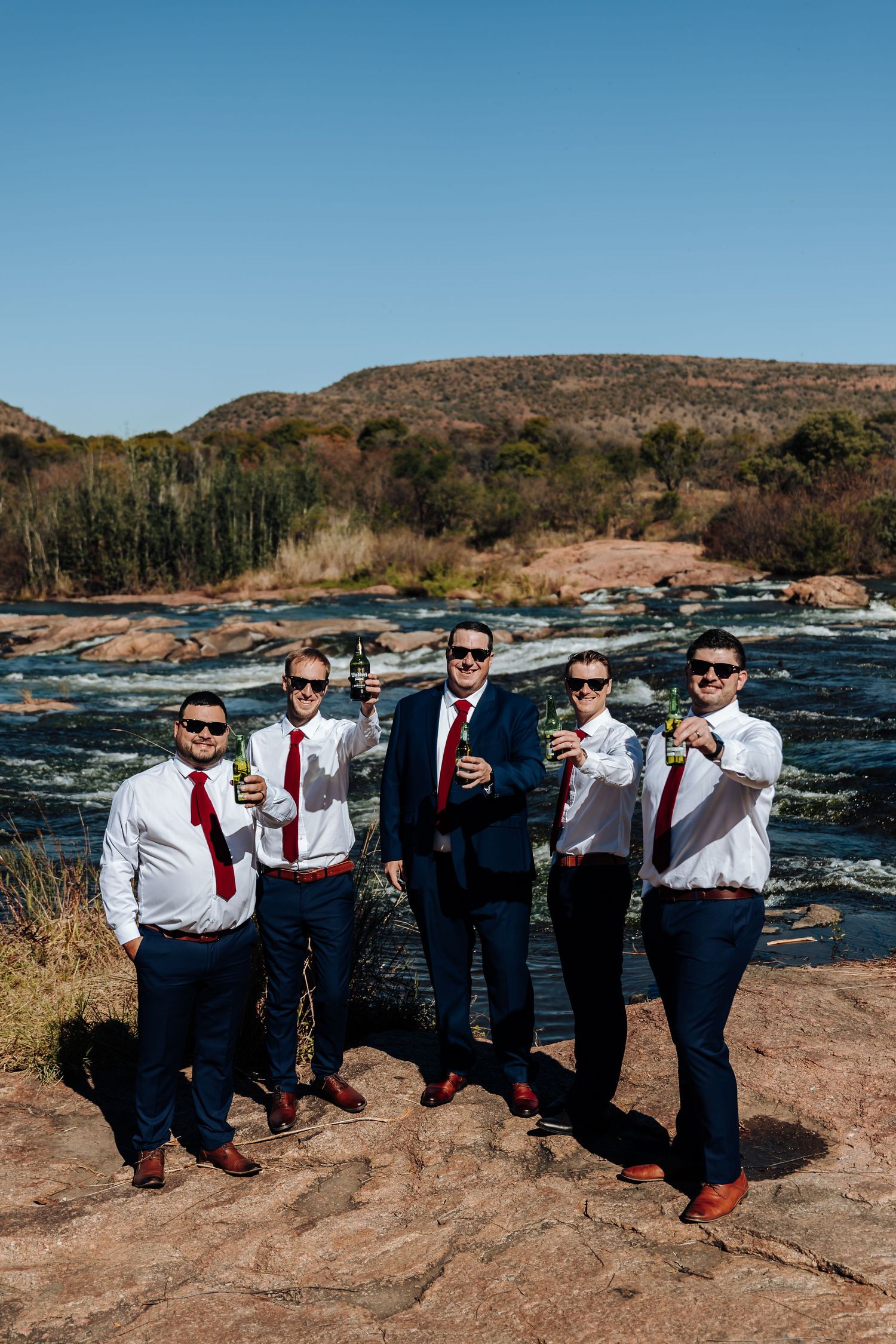 Five groomsmen in navy suits and red ties toasting with beer bottles by a scenic river on a sunny wedding day.