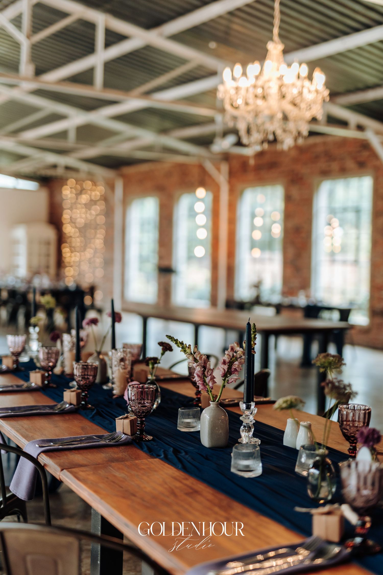 Elegant wedding reception venue with long wooden tables, navy runners, floral centerpieces, and a crystal chandelier.