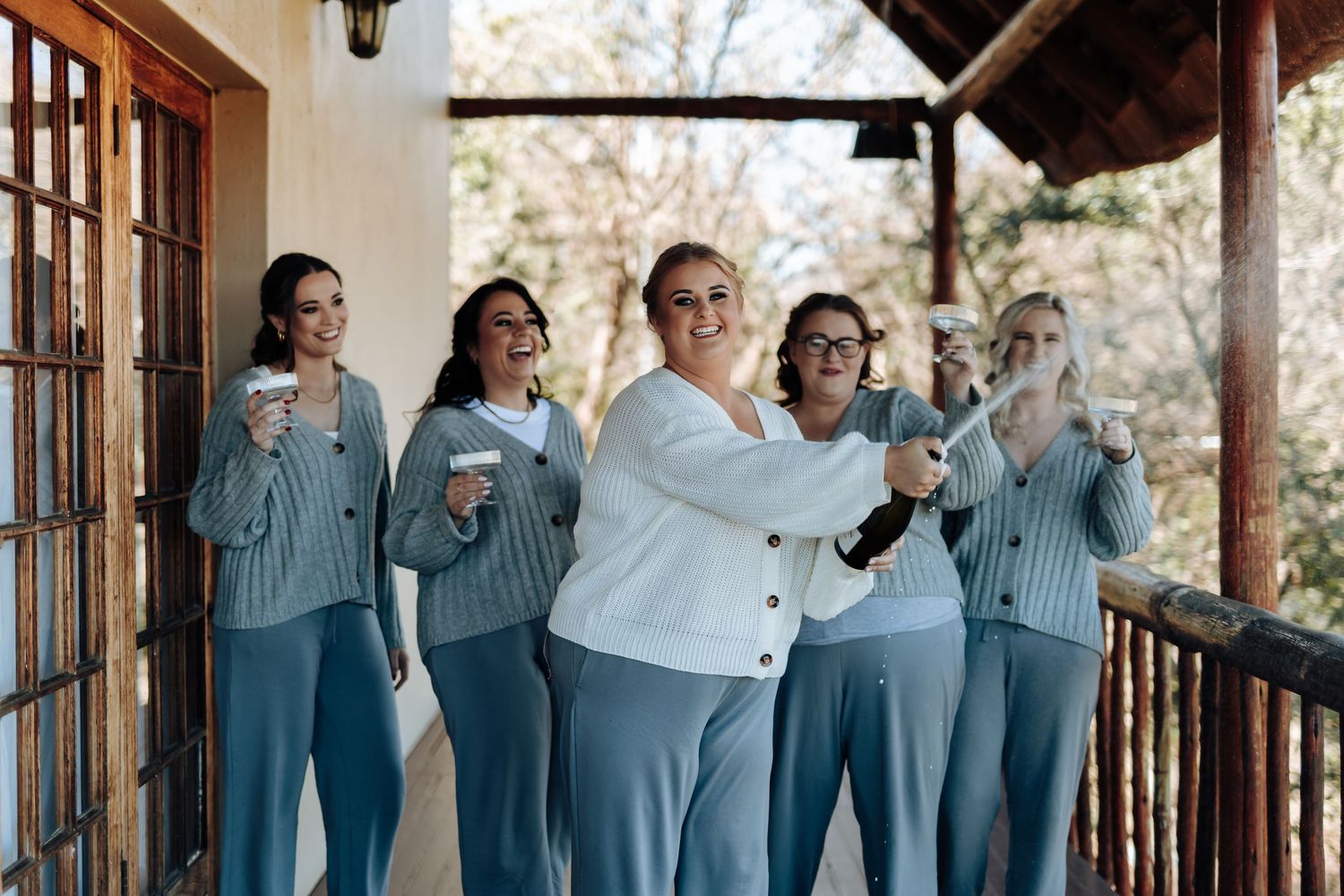 Bride in white cardigan pops champagne with bridesmaids in matching blue-gray outfits on a rustic wooden porch.
