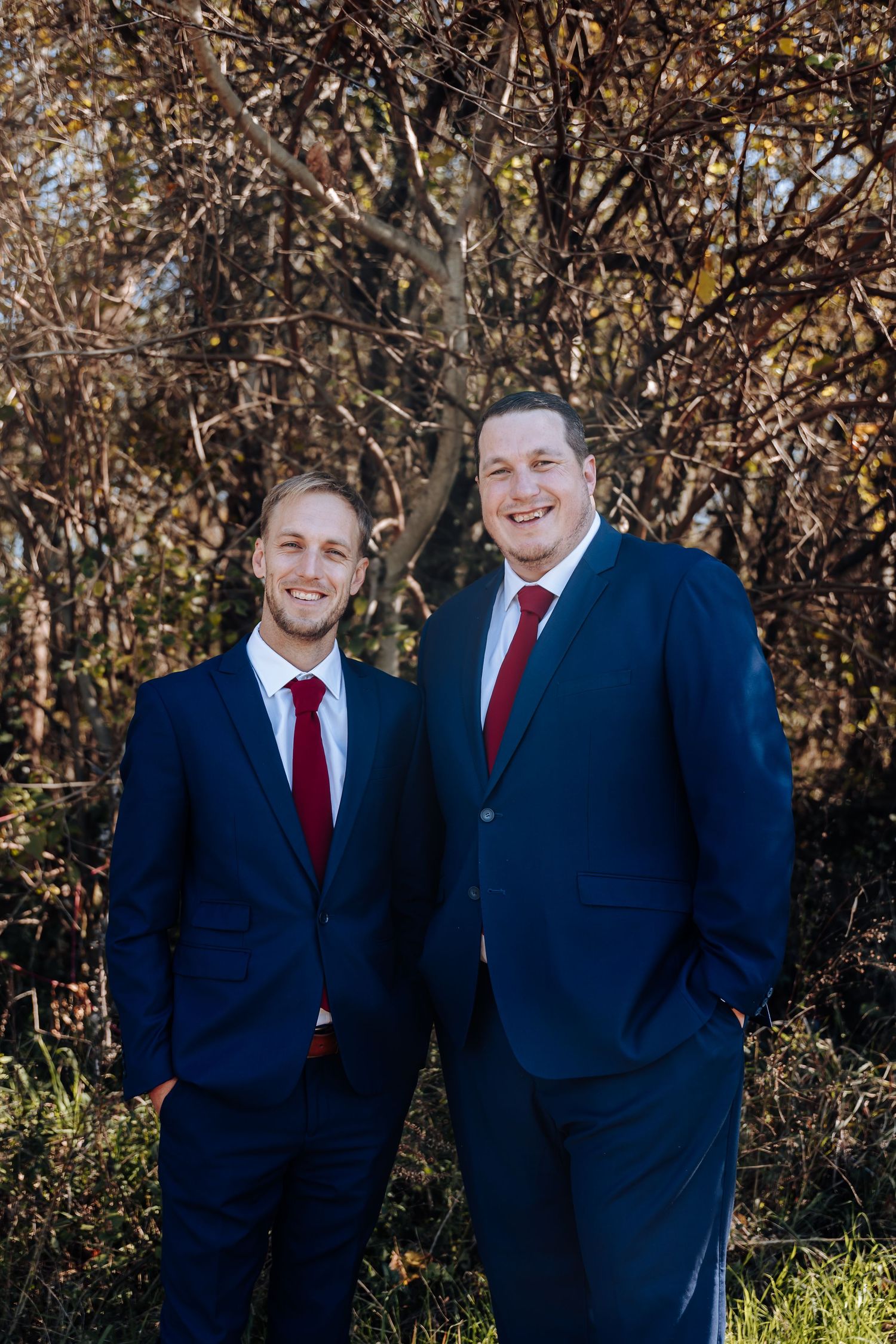Two men in matching navy blue suits and red ties smile outdoors with autumn foliage in the background.