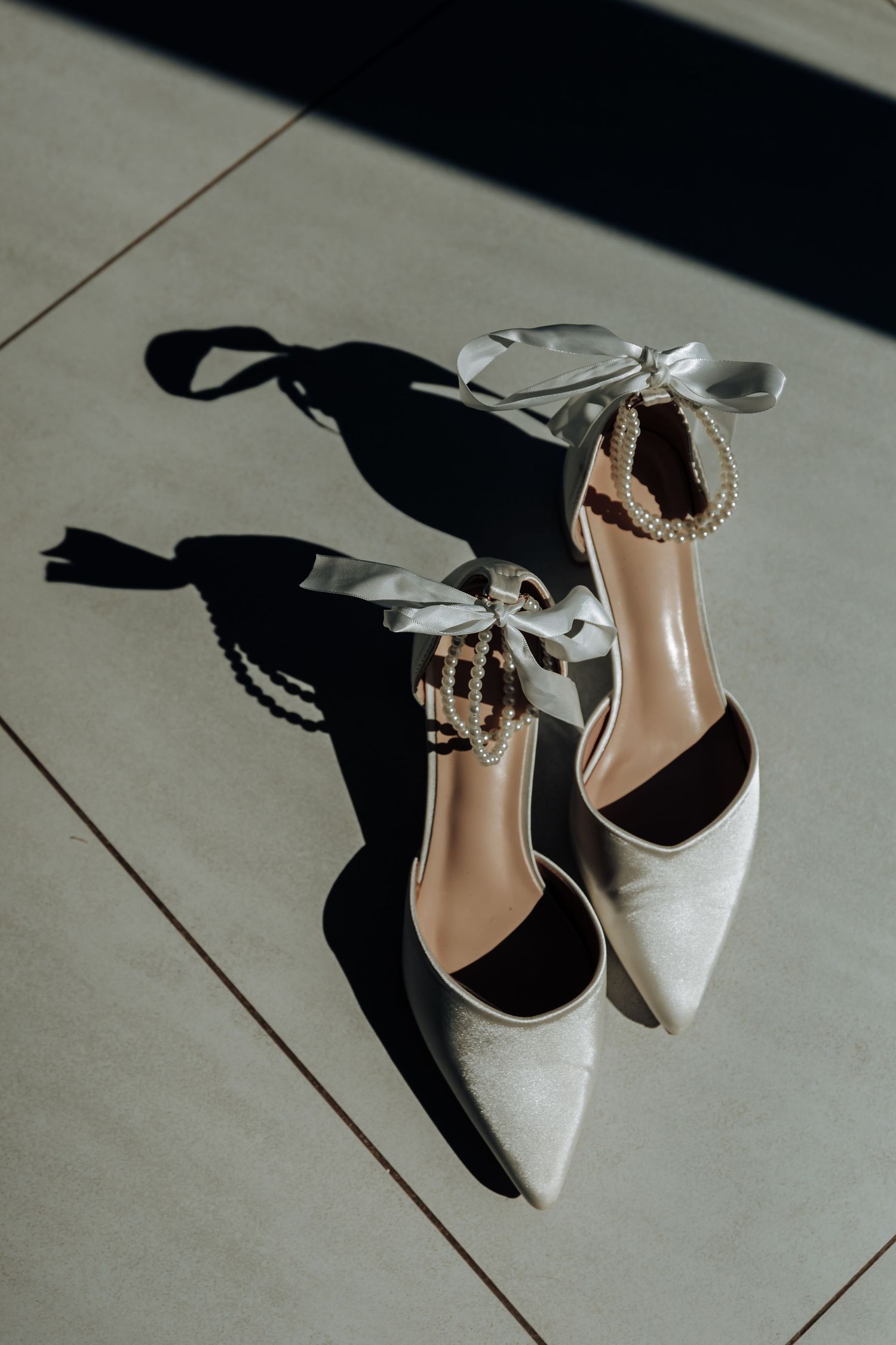 Elegant white satin bridal heels with crystal ankle straps and ribbon ties casting shadows on gray tiles.