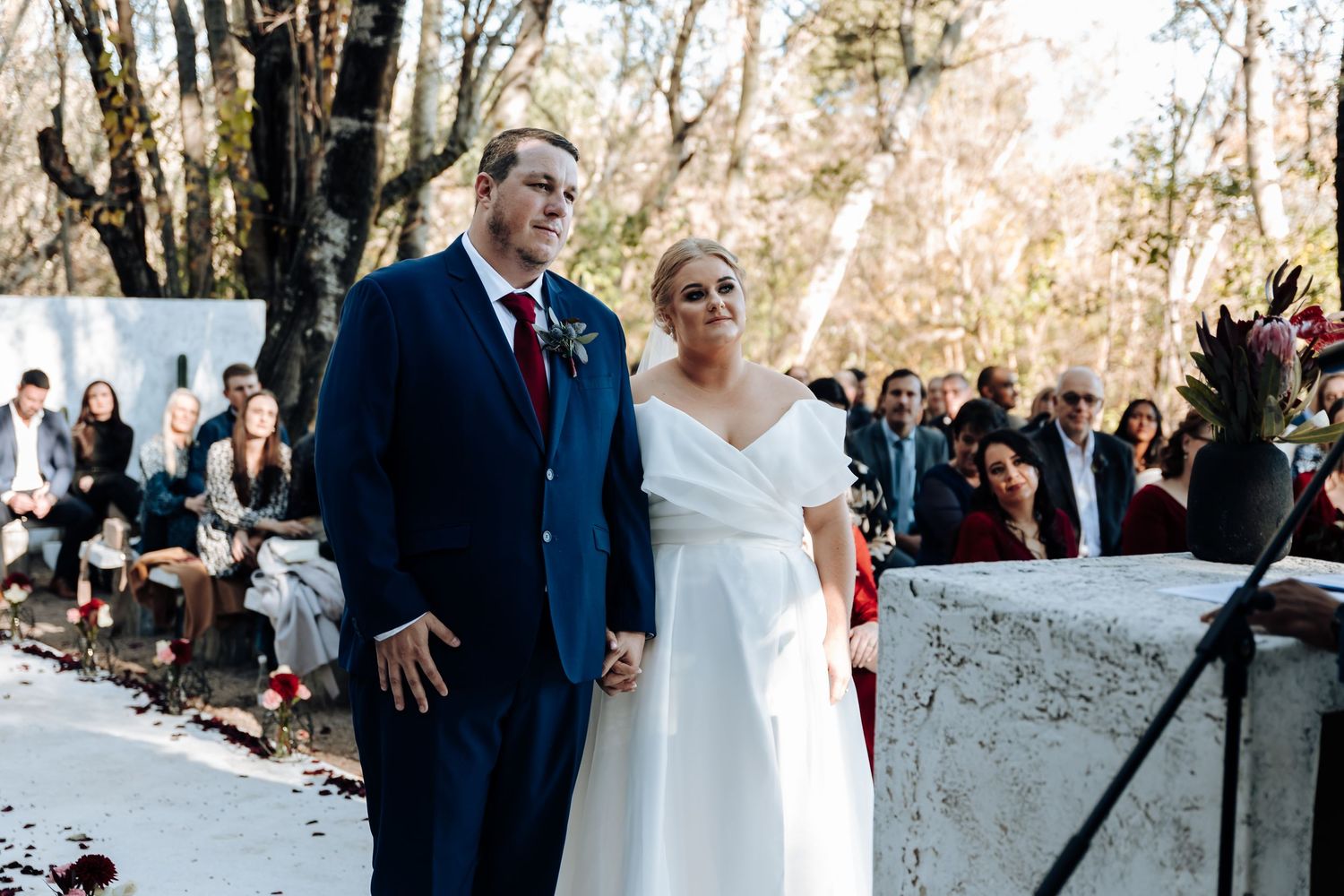 Bride in white off-shoulder gown and groom in navy suit hold hands smiling at outdoor wedding ceremony.