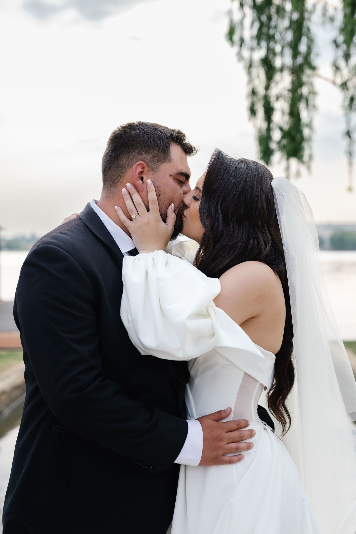 A romantic wedding moment captured outdoors with a couple sharing a kiss under willow trees by the water.