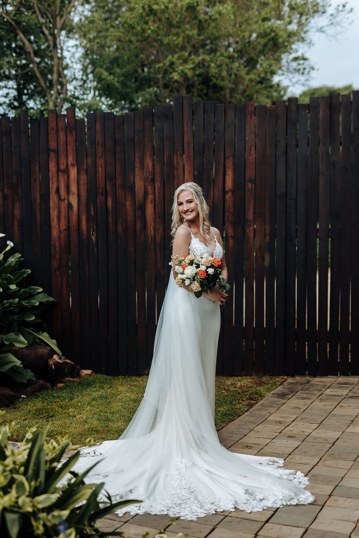 A bride in a flowing white lace wedding gown stands in front of a wooden fence on a stone patio.