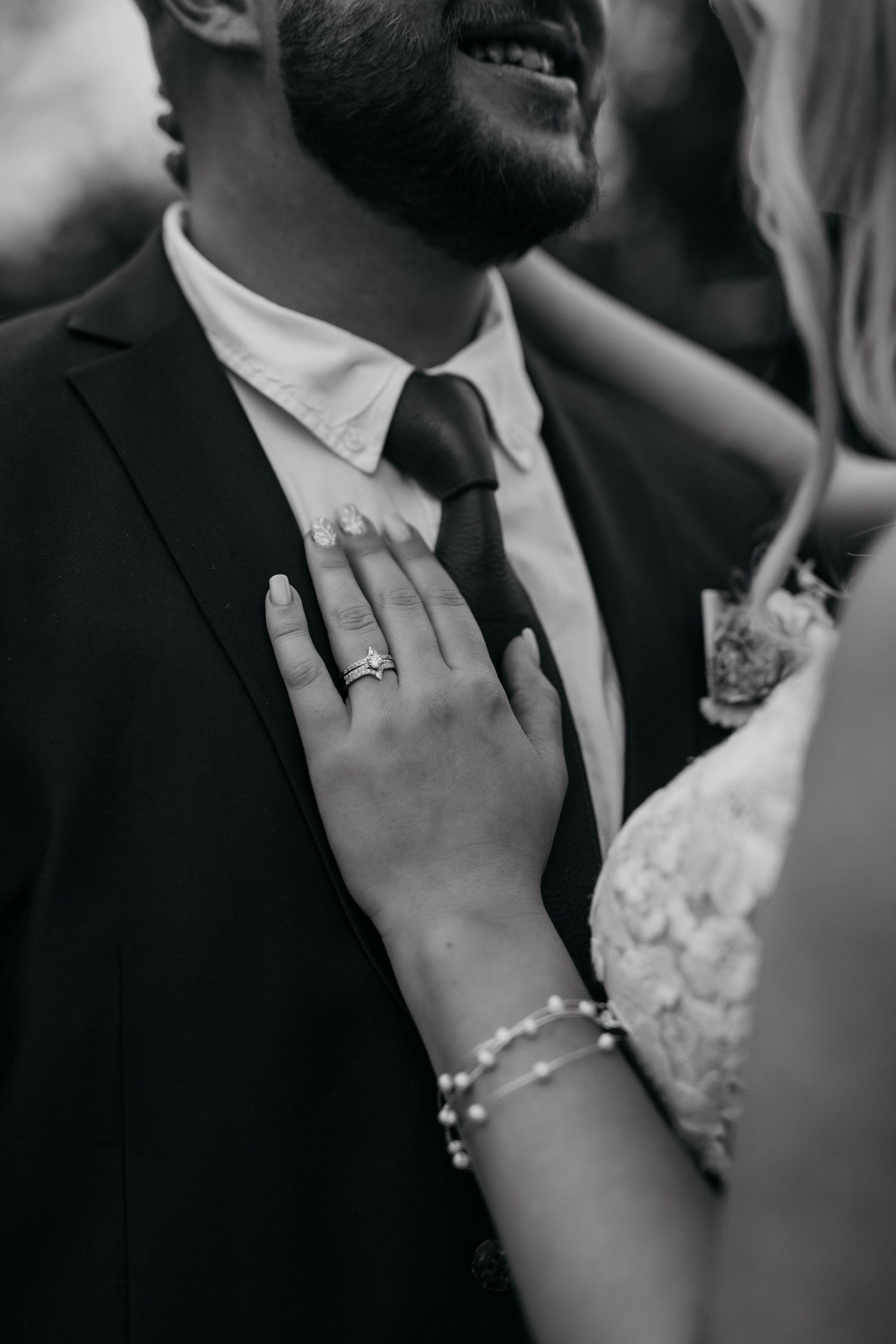 Black and white wedding photo showing a close-up of hands with rings and jewelry against formal attire.