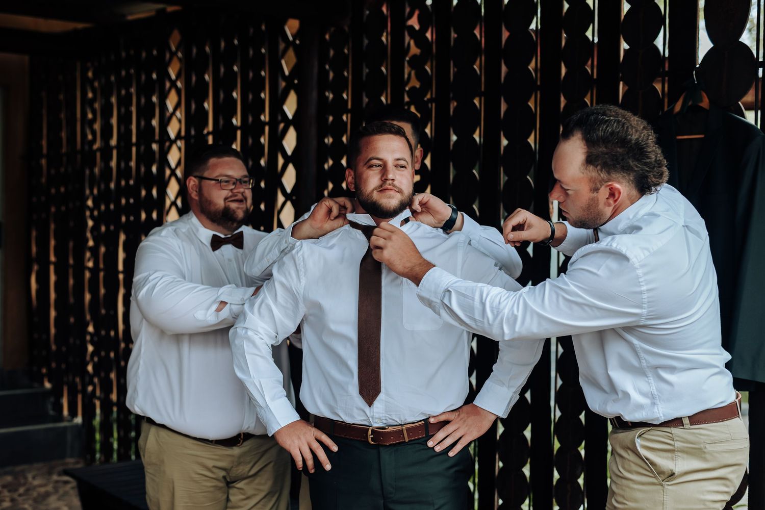 Groomsmen help adjust a brown tie on a man in a white dress shirt before a wedding ceremony.