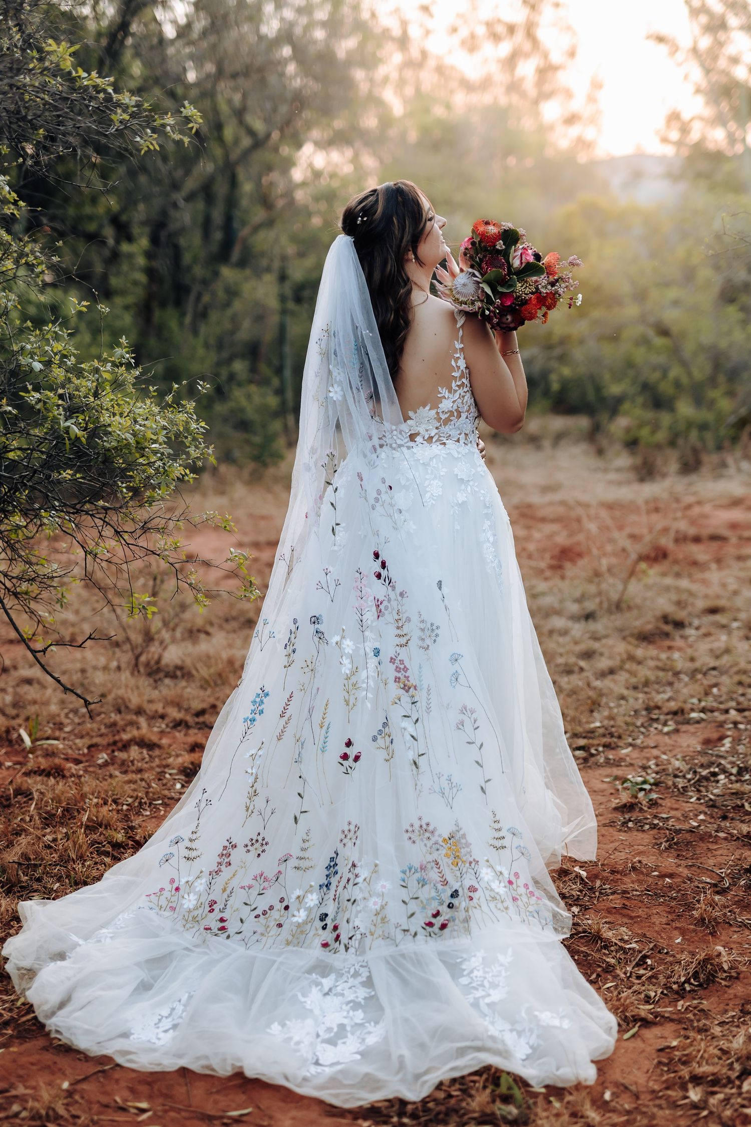 A flowing white lace wedding dress with a long veil photographed from behind in a rustic outdoor autumn setting at sunset.