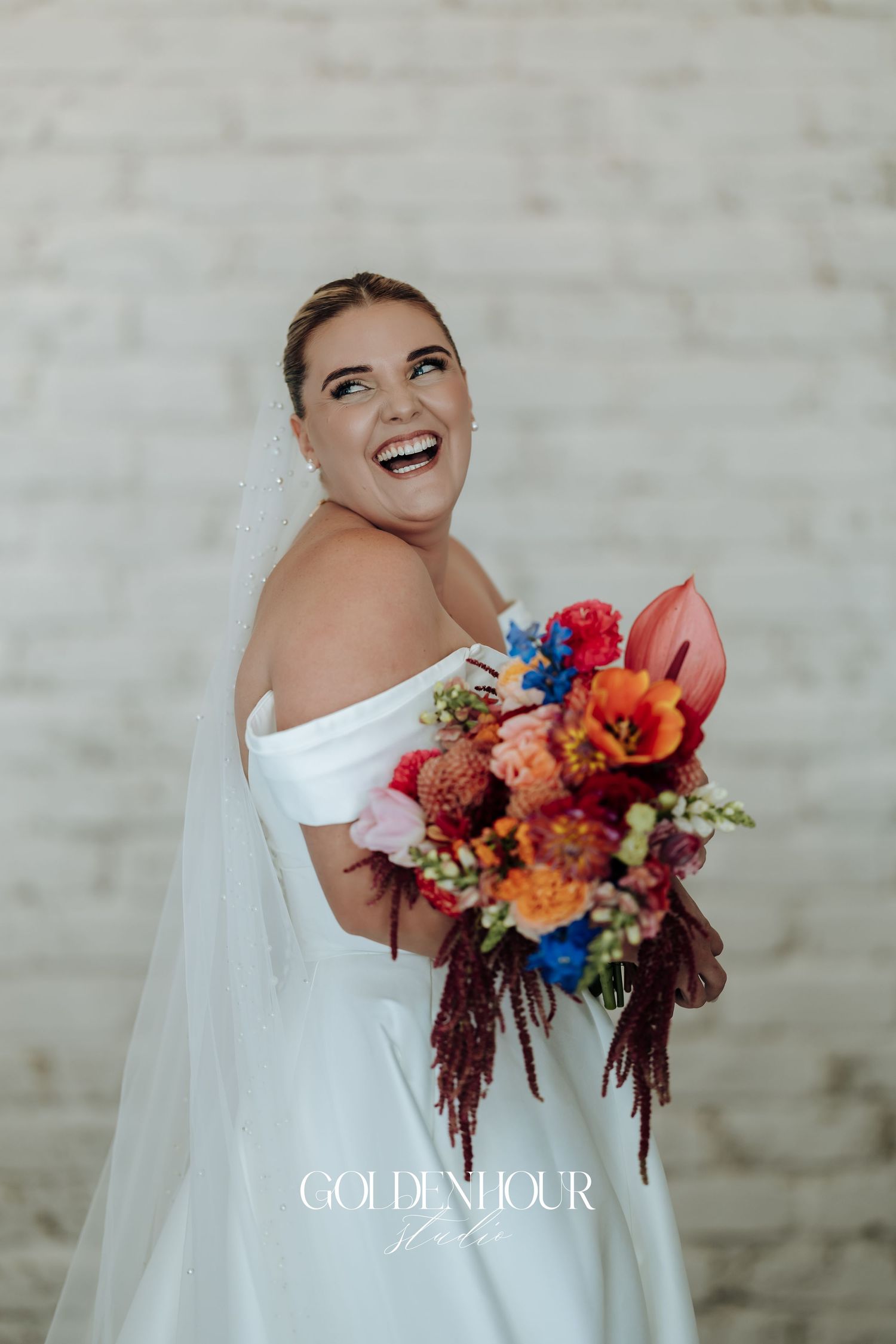 Joyful bride in off-shoulder wedding dress laughs while holding vibrant autumn bouquet with red and blue flowers.