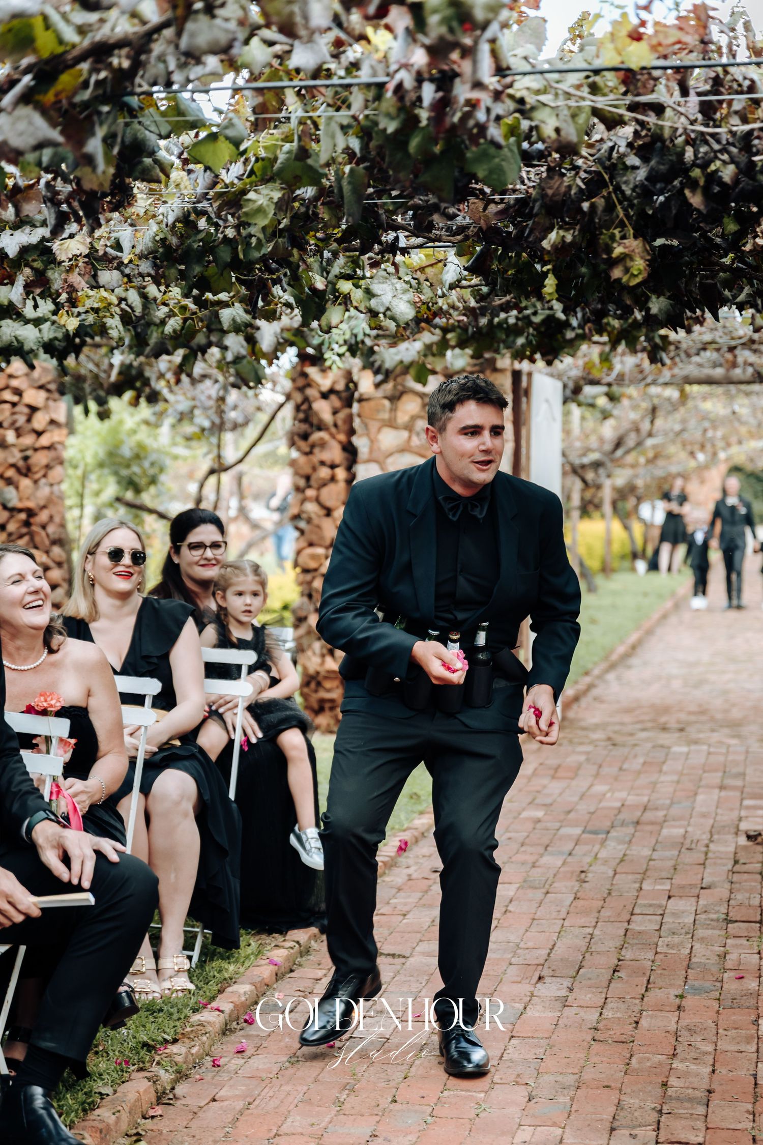 Wedding party members in black attire share a fun candid moment on brick pathway during outdoor celebration.