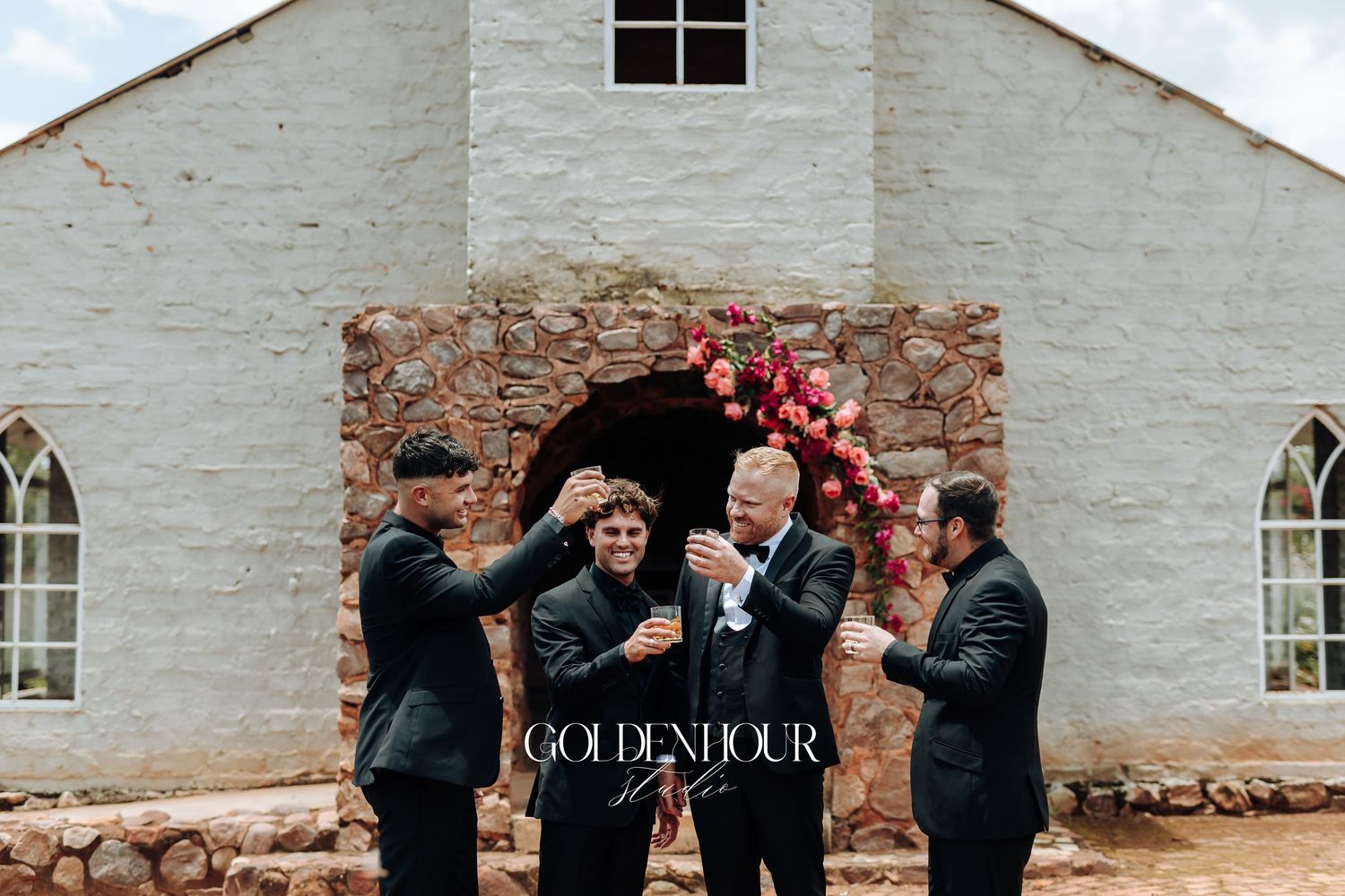 Wedding party celebrates joyfully outside historic white stone building on sunny day.