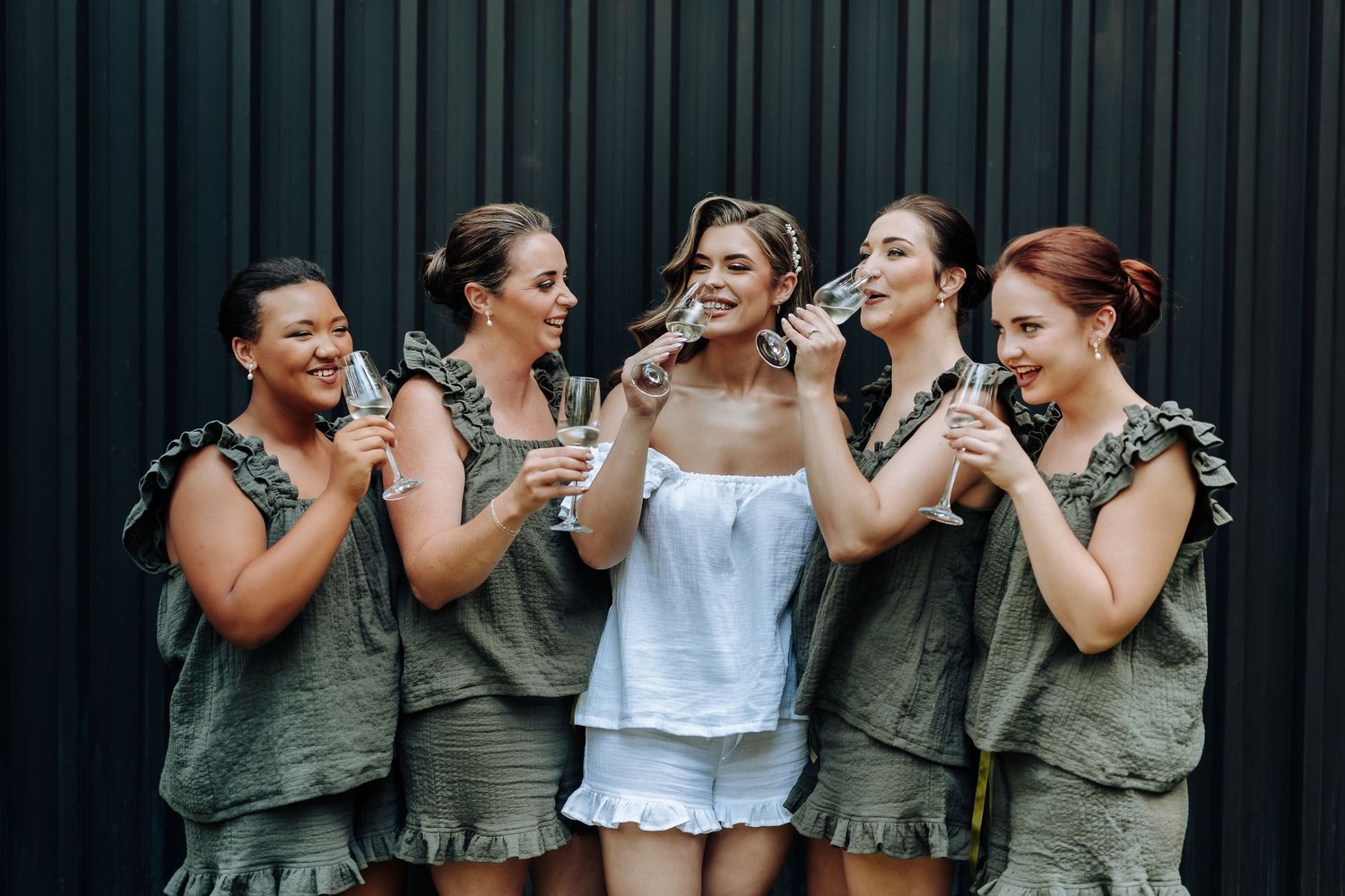 Wedding party in coordinated outfits sharing drinks and laughter against a dark backdrop.