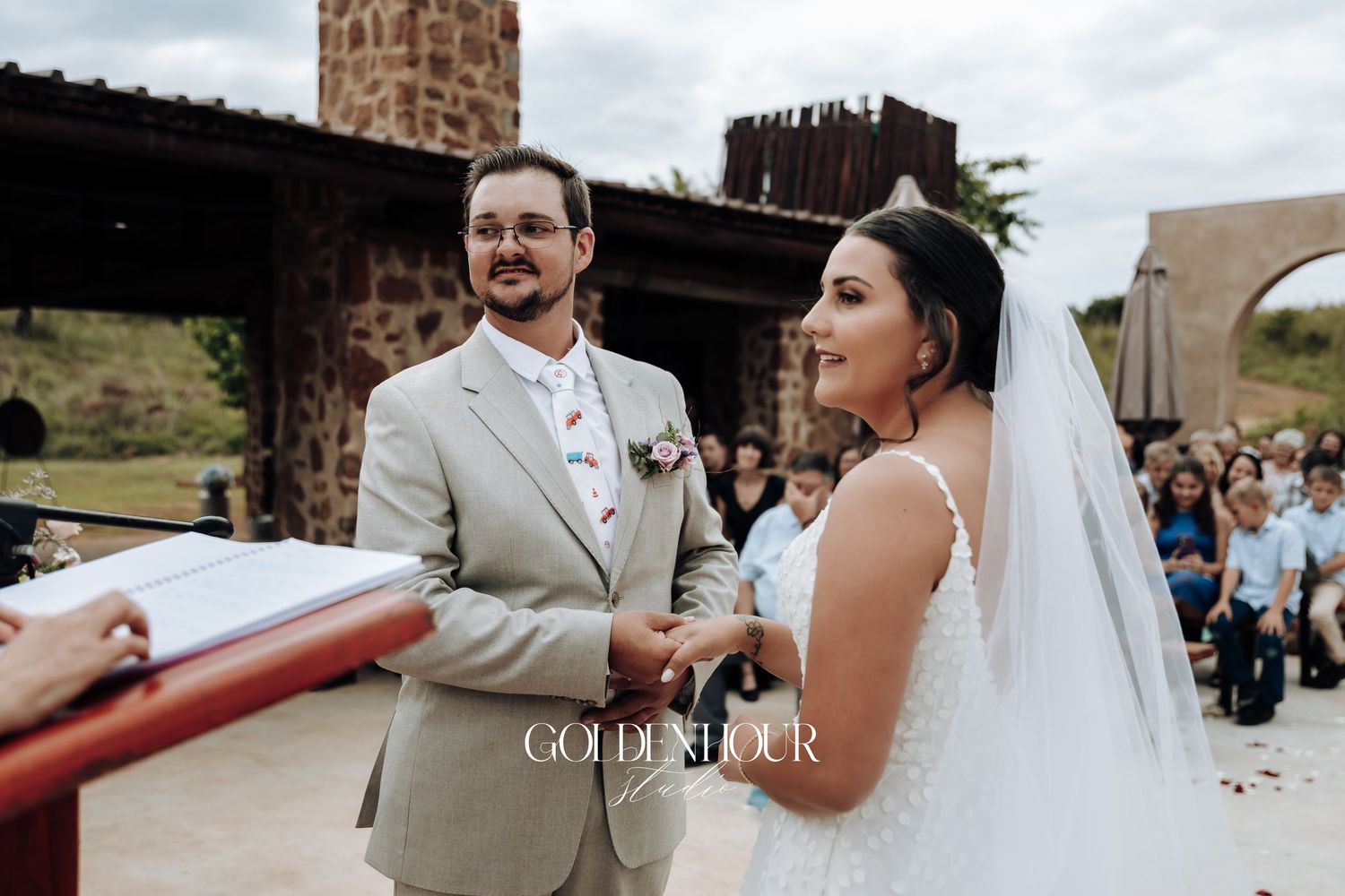 Wedding ceremony moment under rustic stone archway with guests in background.