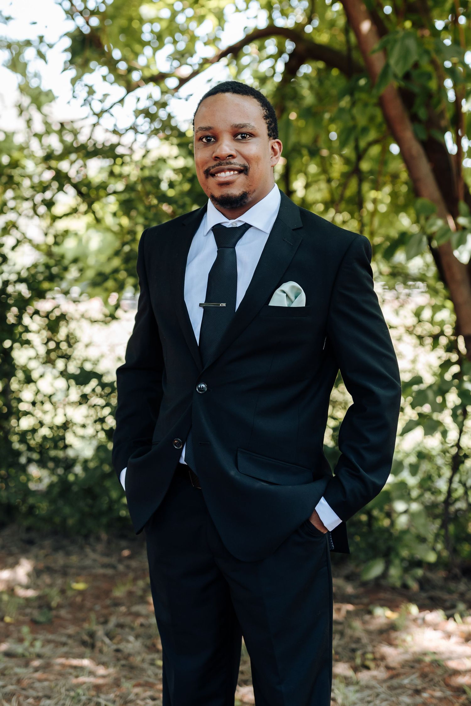 Groomsmen in black suits and ties posing casually outdoors with trees in background.