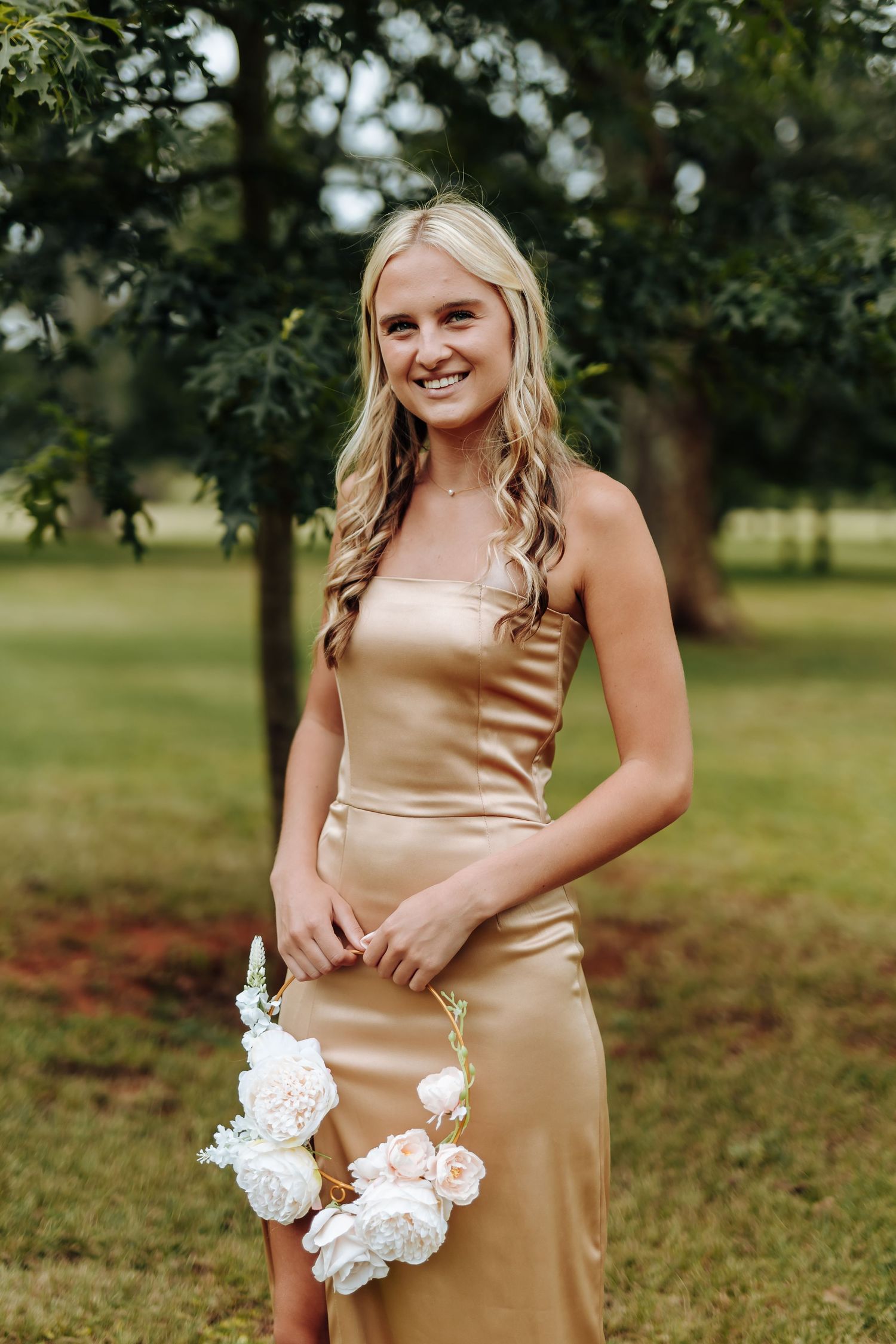 A woman in a champagne silk dress poses outdoors holding white flowers in a grassy field.