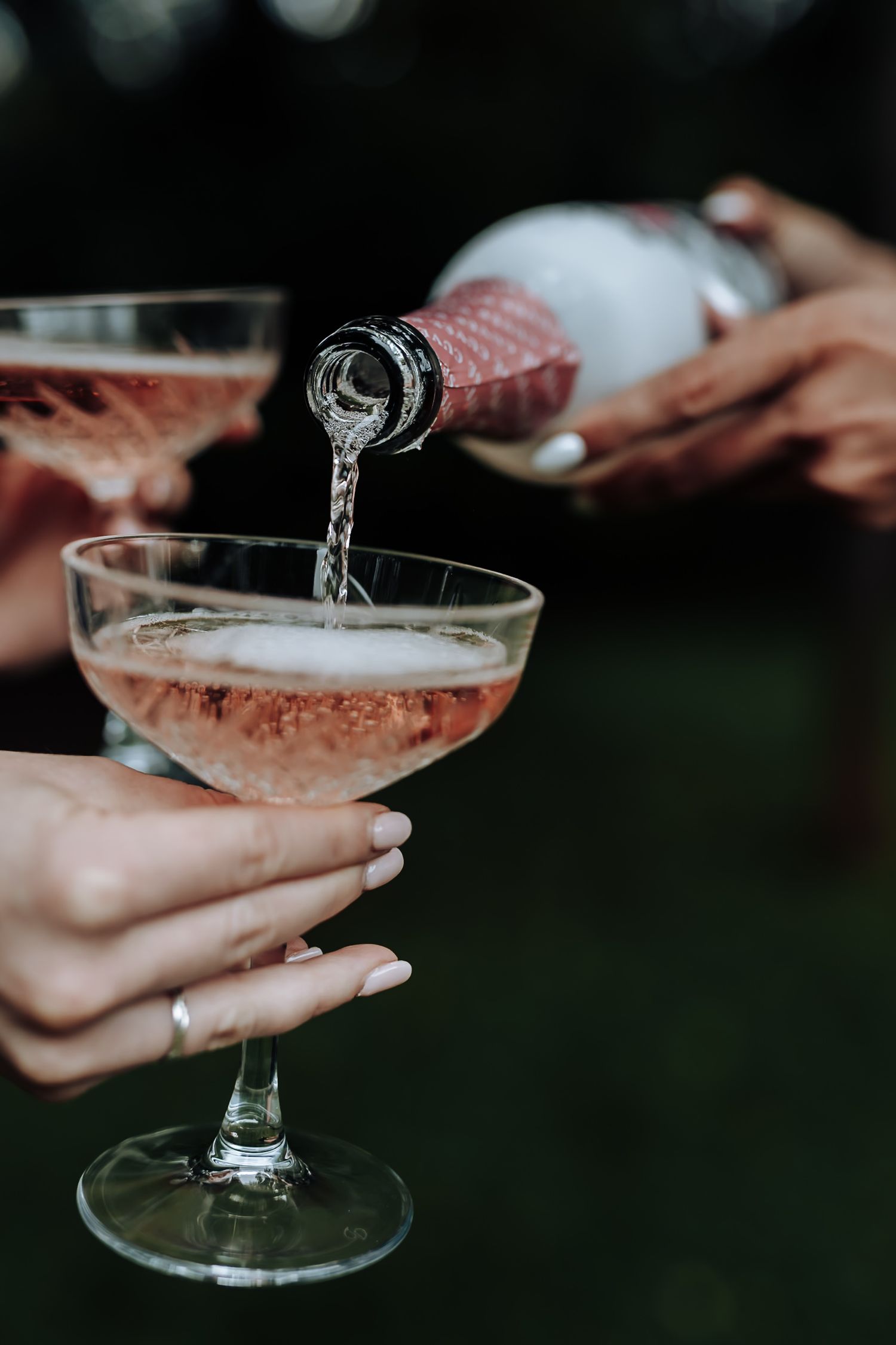 Hands holding elegant coupe champagne glasses filled with sparkling pink cocktails against a dark background.
