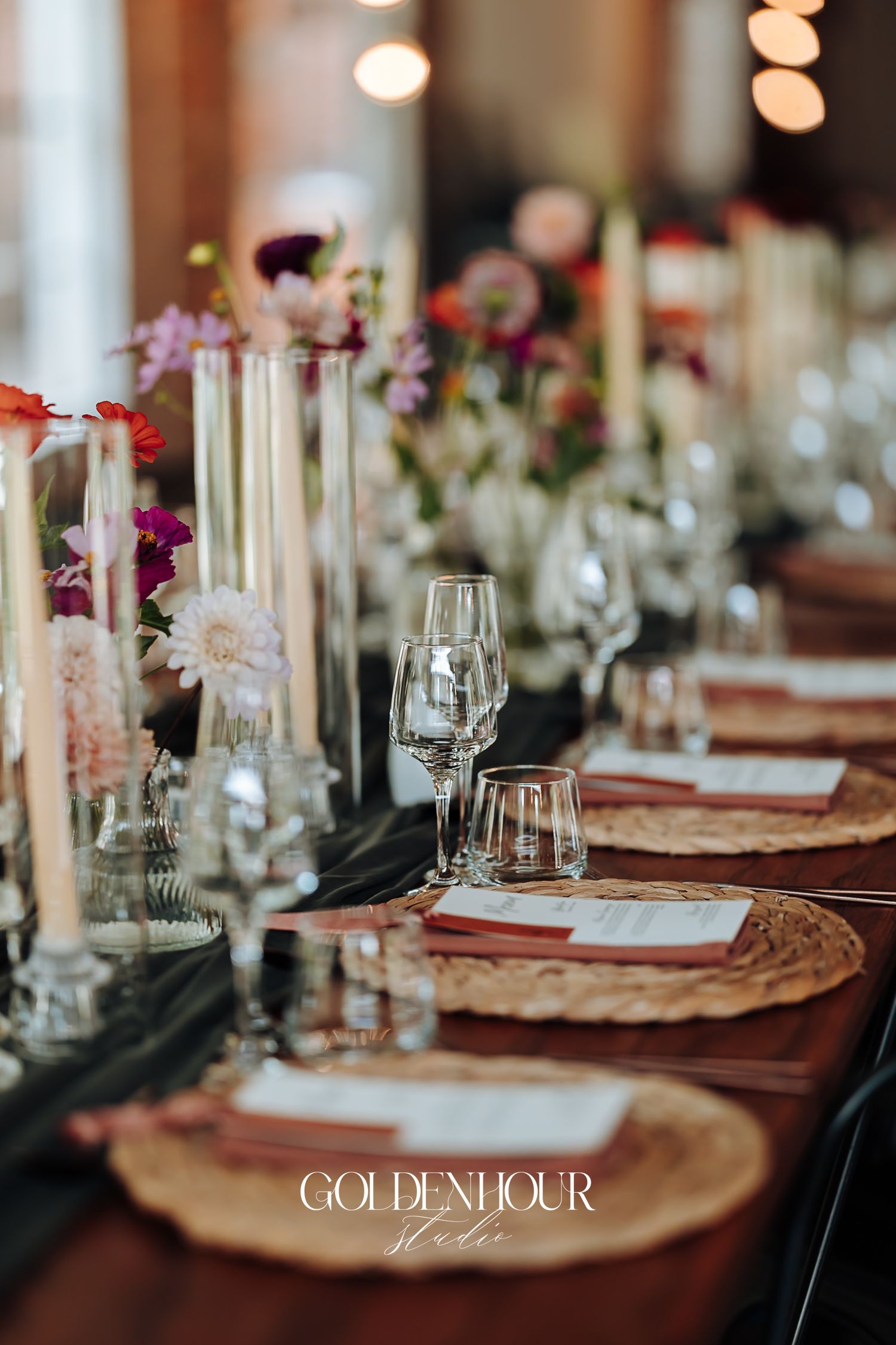 Elegant wedding reception table with woven chargers, crystal glassware, taper candles, and colorful floral centerpieces.