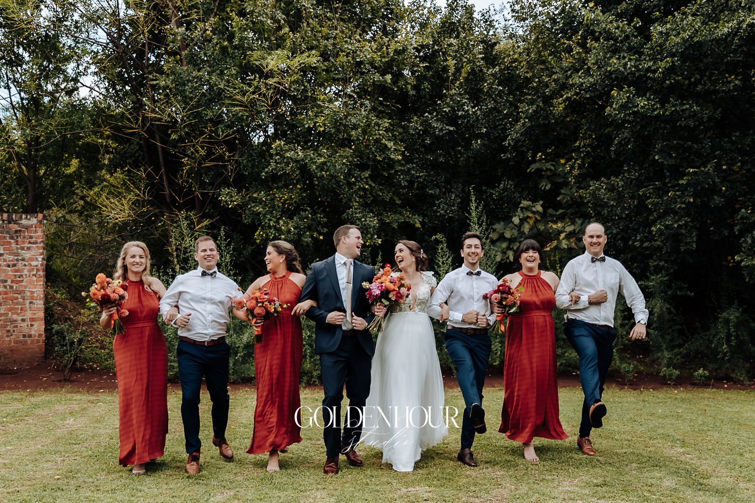 Wedding party in rust bridesmaid dresses and suits joyfully running on lawn with bouquets outdoors.