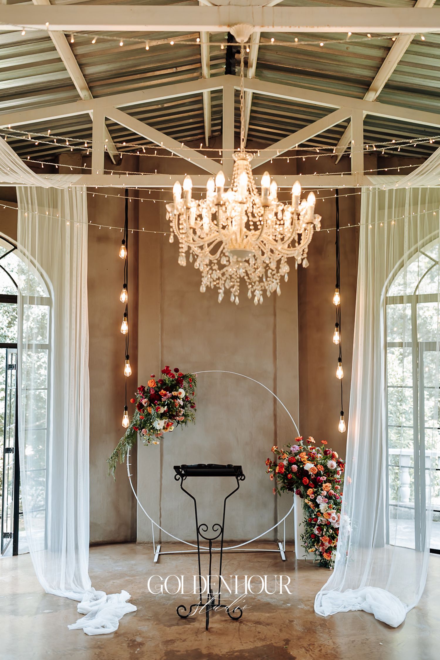 Elegant wedding ceremony setup with crystal chandelier, white draping, circular arch, and colorful floral arrangements.