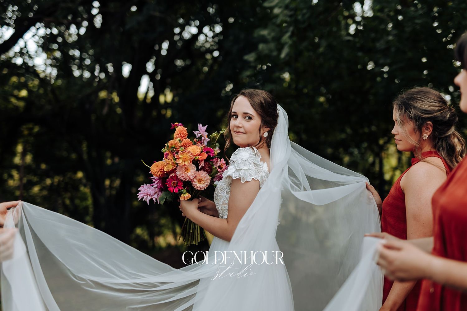 Bride in lace gown and veil holding colorful bouquet looking back with bridesmaid in red.
