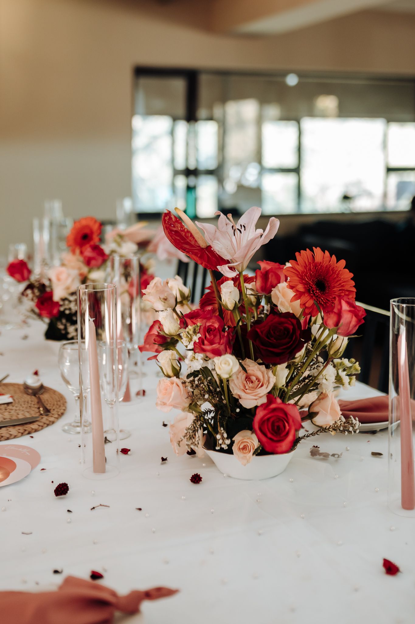 Detailed shot of wedding table decor with red and pink flowers, tall candles, and scattered rose petals.