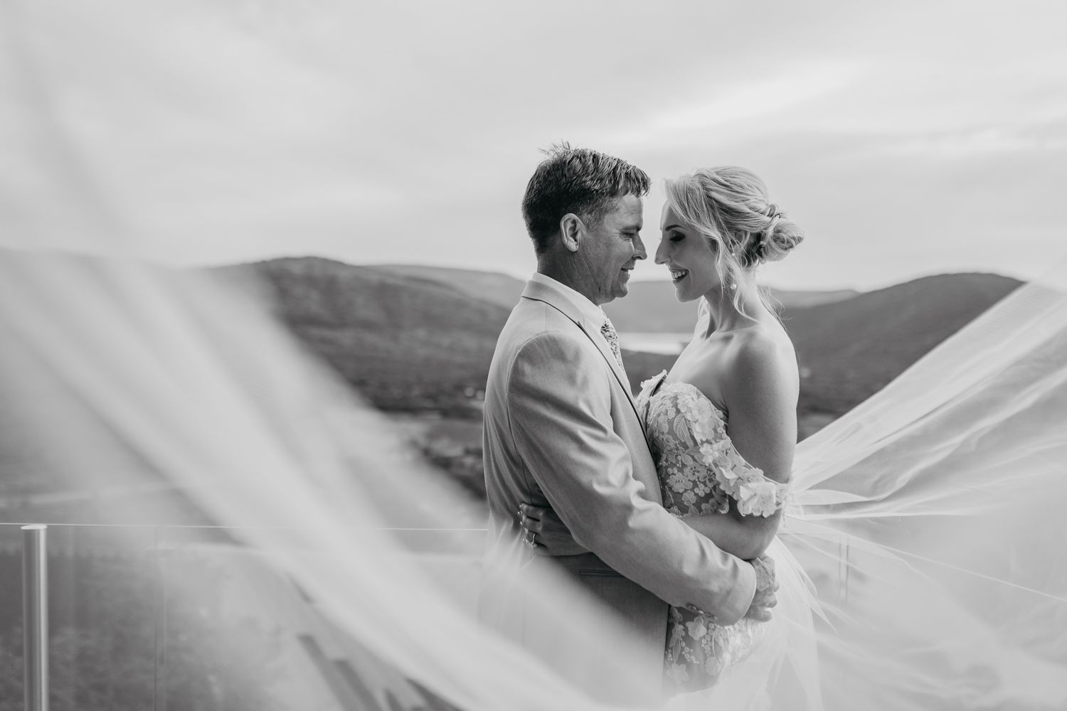 Black and white wedding photo with veil flowing in the wind against a mountain backdrop.
