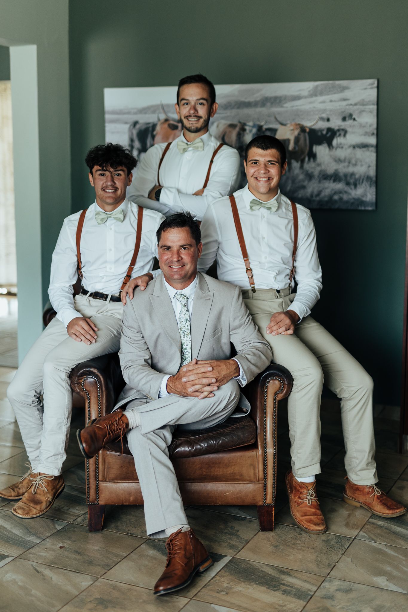 Wedding party poses together with suspenders and light colored suits on leather furniture.