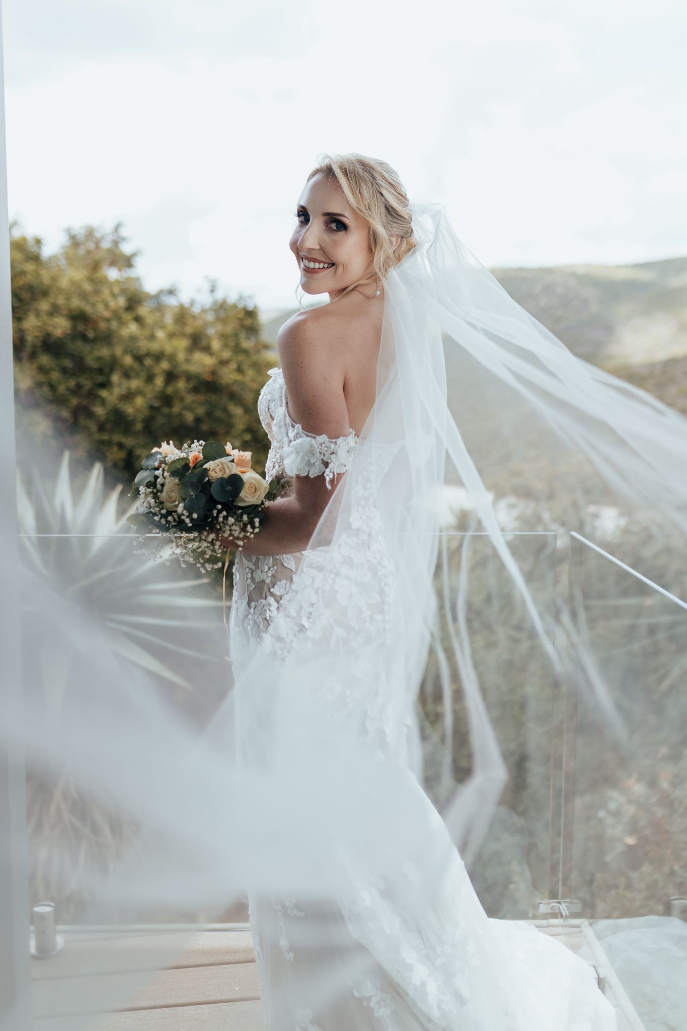 Bride with flowing veil and fitted lace dress smiles while holding bouquet on outdoor deck.