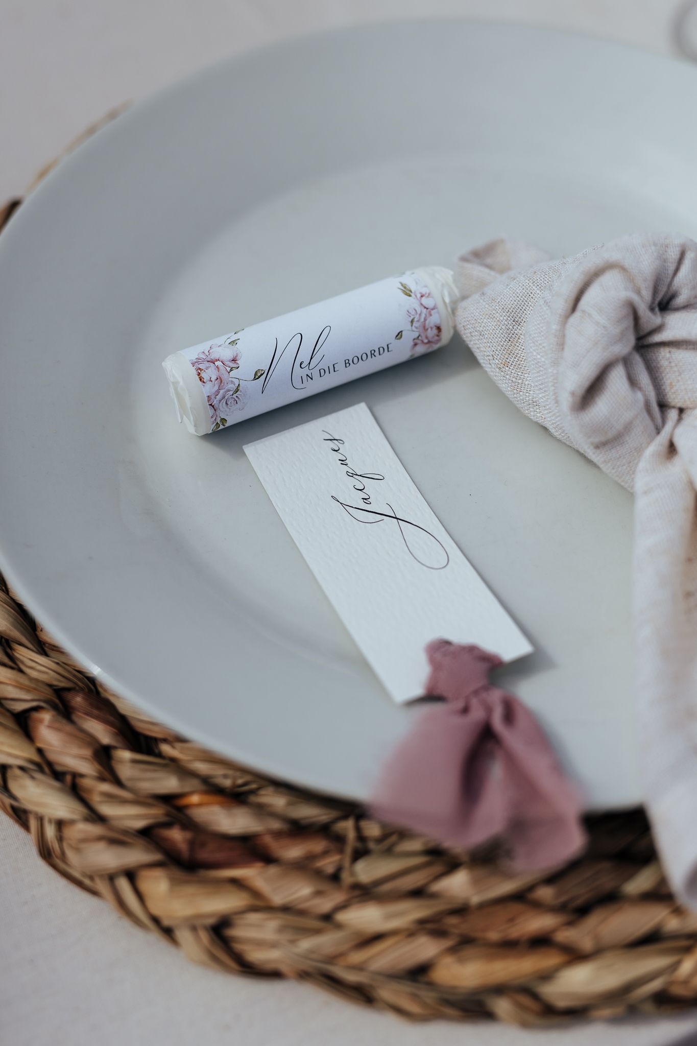 Close-up of place card and rolled menu on white plate with woven charger and pink accent.