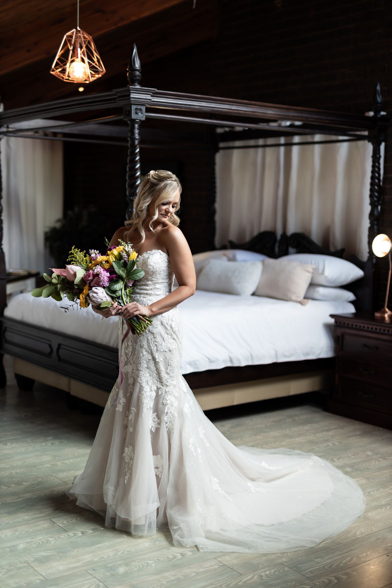 Elegant bride in a fitted lace wedding dress poses with colorful bouquet by a canopy bed.