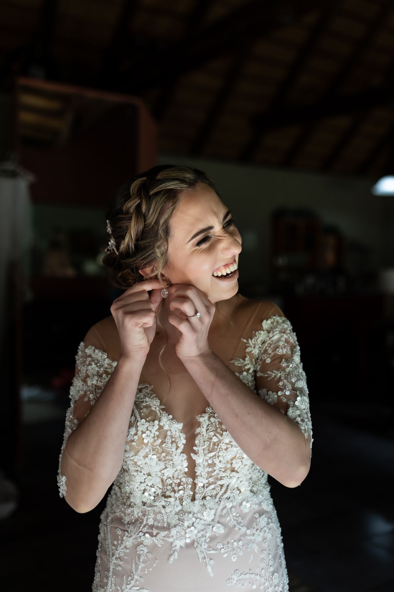 A laughing bride in a beaded lace wedding dress putting on earrings during wedding preparations.