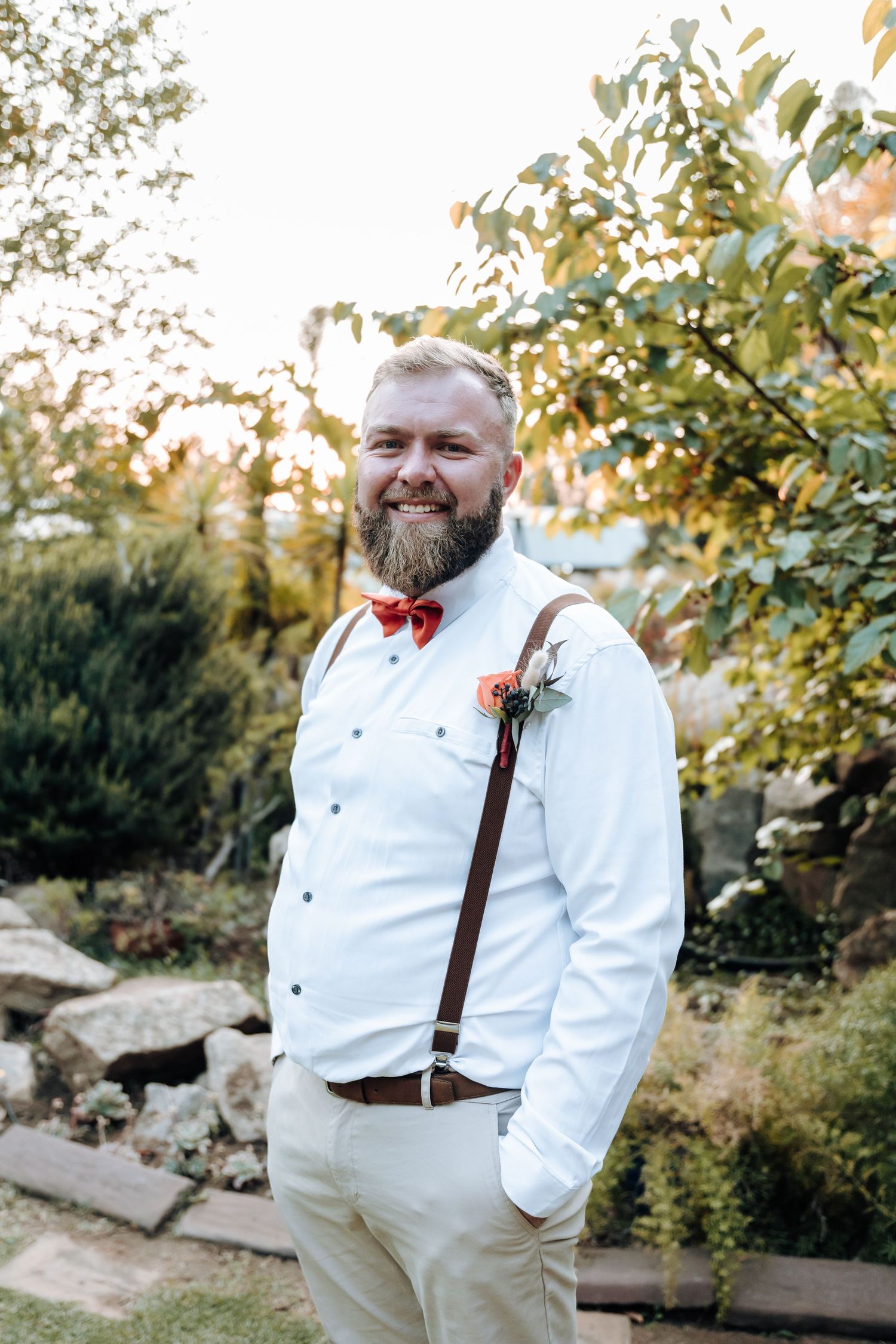Groomsman in white shirt with red bow tie and suspenders poses in garden setting.