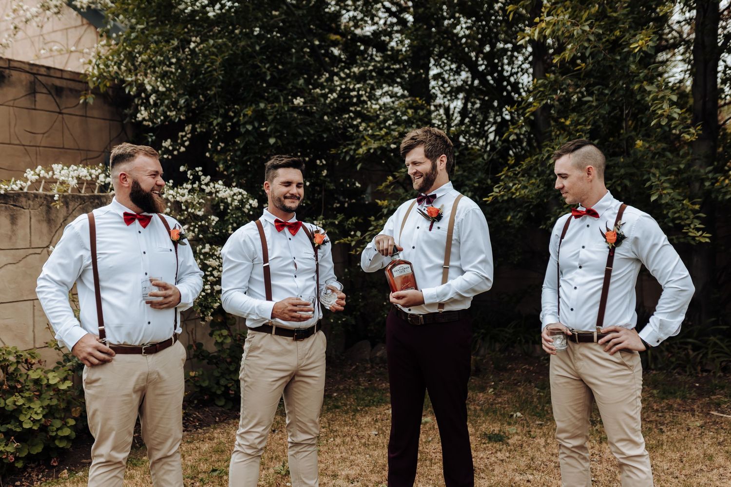 Groomsmen in suspenders and bow ties pose casually outdoors in matching attire.