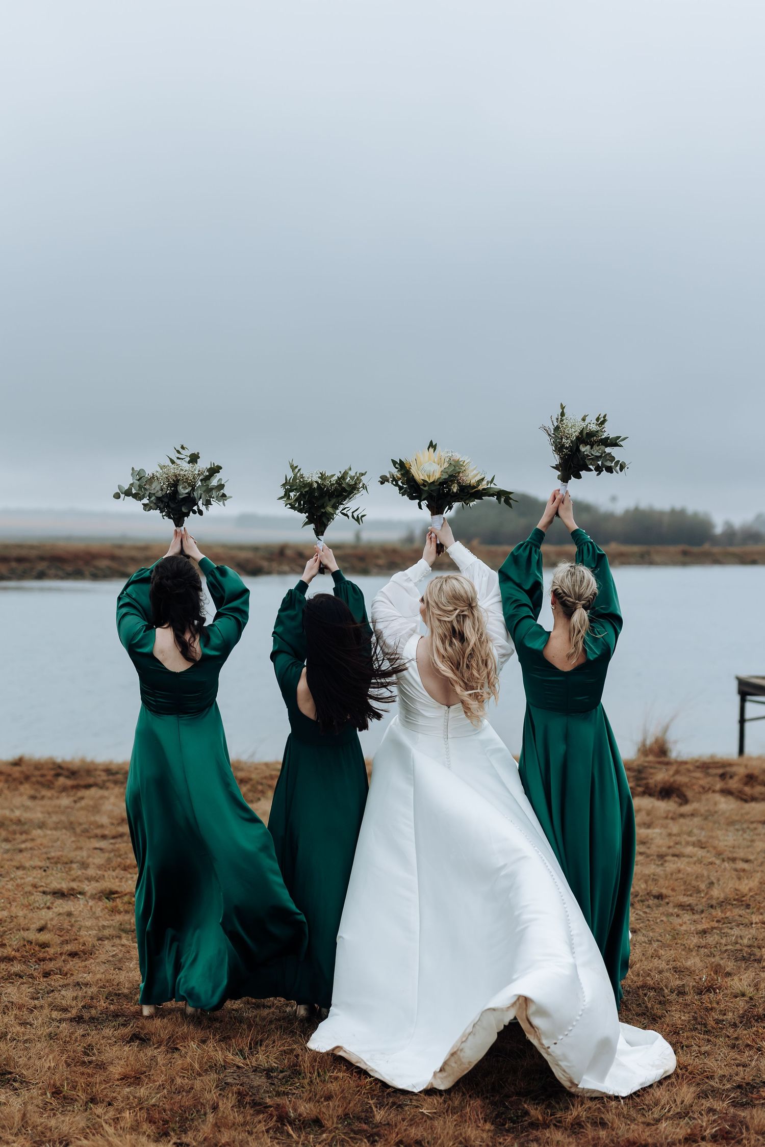 Bridesmaids in emerald gowns hold bouquets up while facing away from camera by the water.